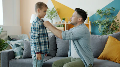 Father buttons son's plaid shirt while sitting on couch