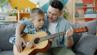 Father teaching son to play guitar on couch.