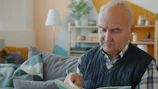 Elderly man reading a book on a couch.