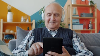 Elderly man with headphones uses a tablet on couch.