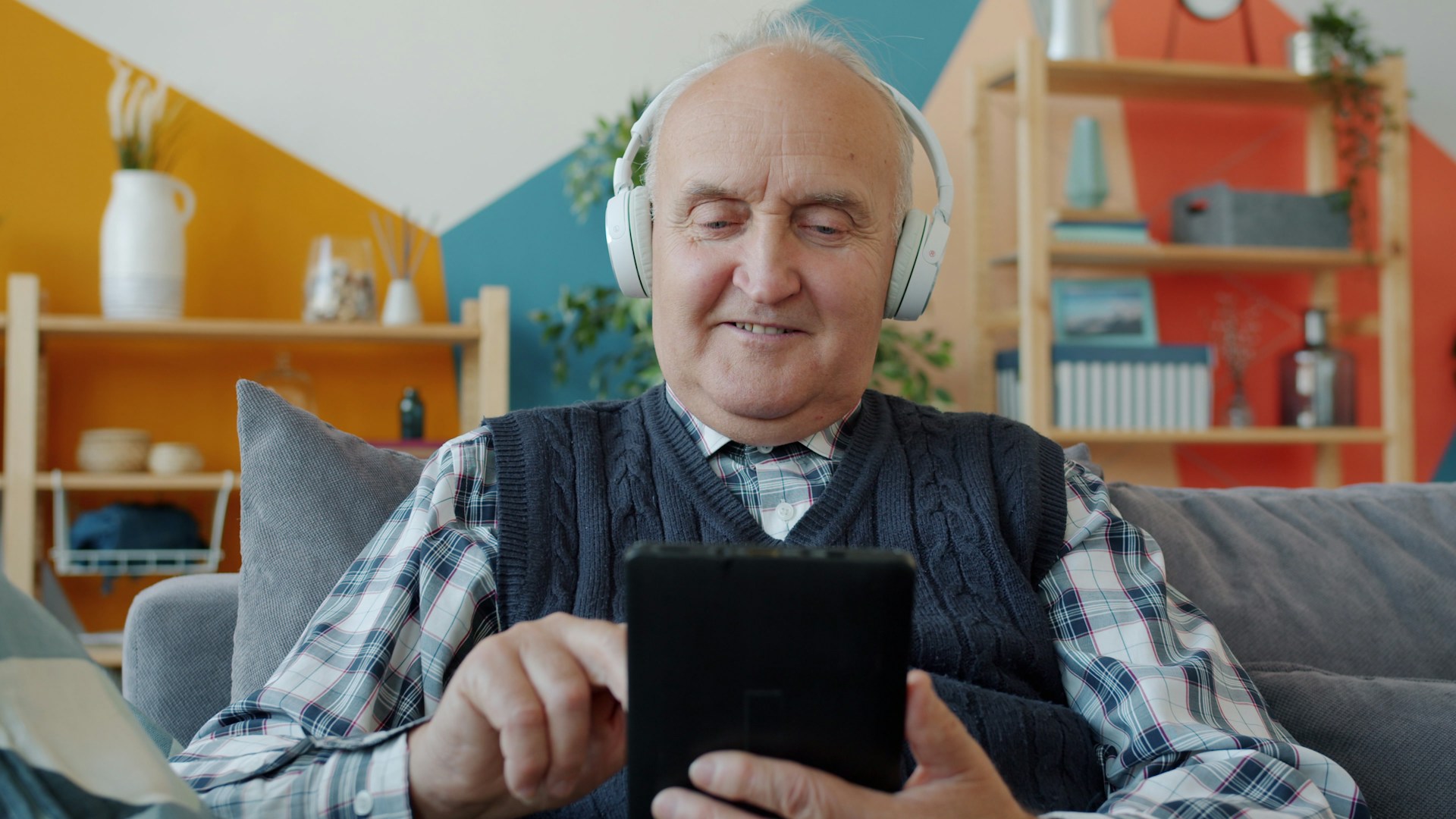 Elderly man with headphones uses a tablet on couch.