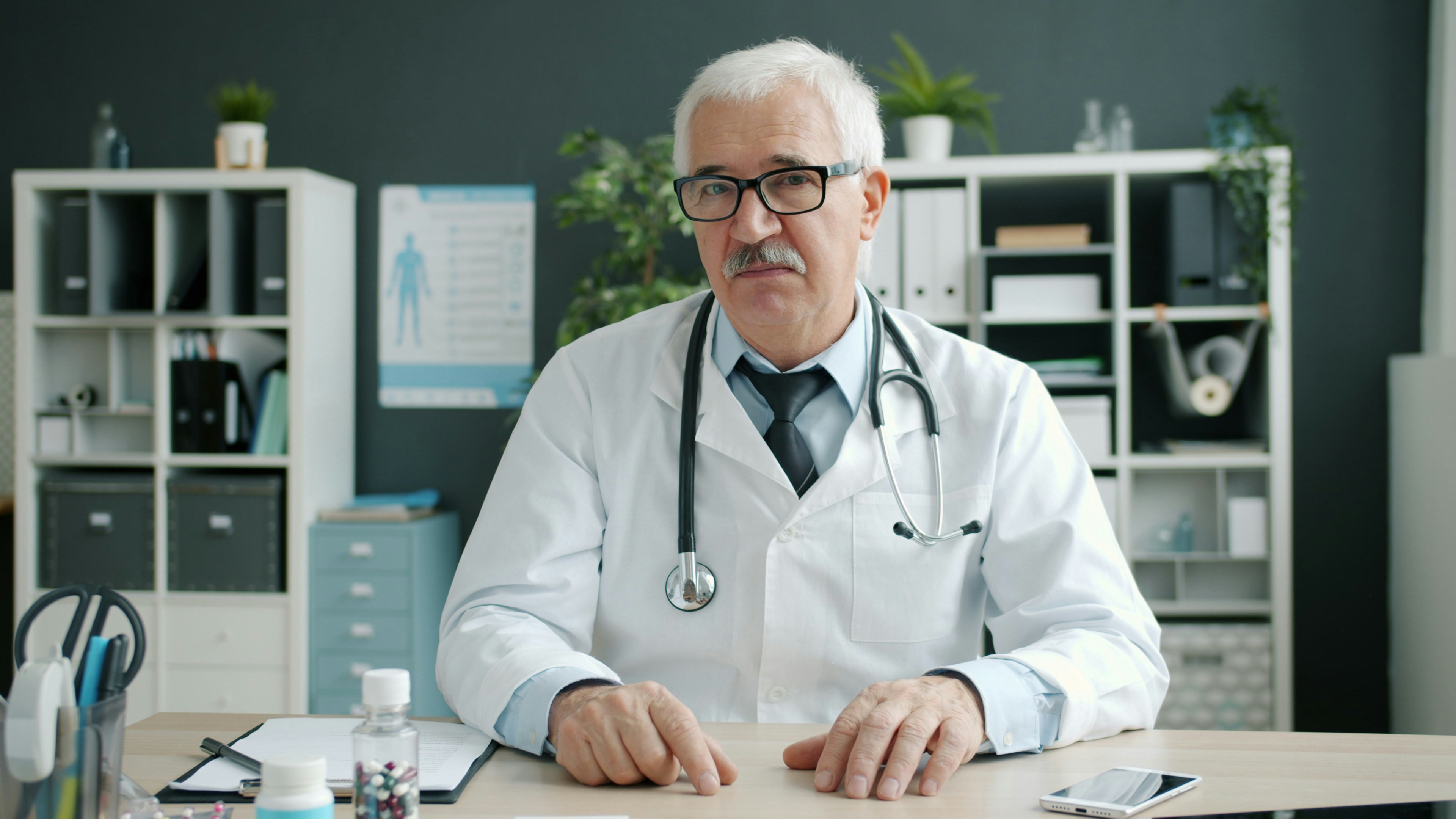 Portrait of joyful doctor senior man in uniform talking waving hand showing bottles with pills in hospital office during online call with patient. Medicine, people and consultation concept.
