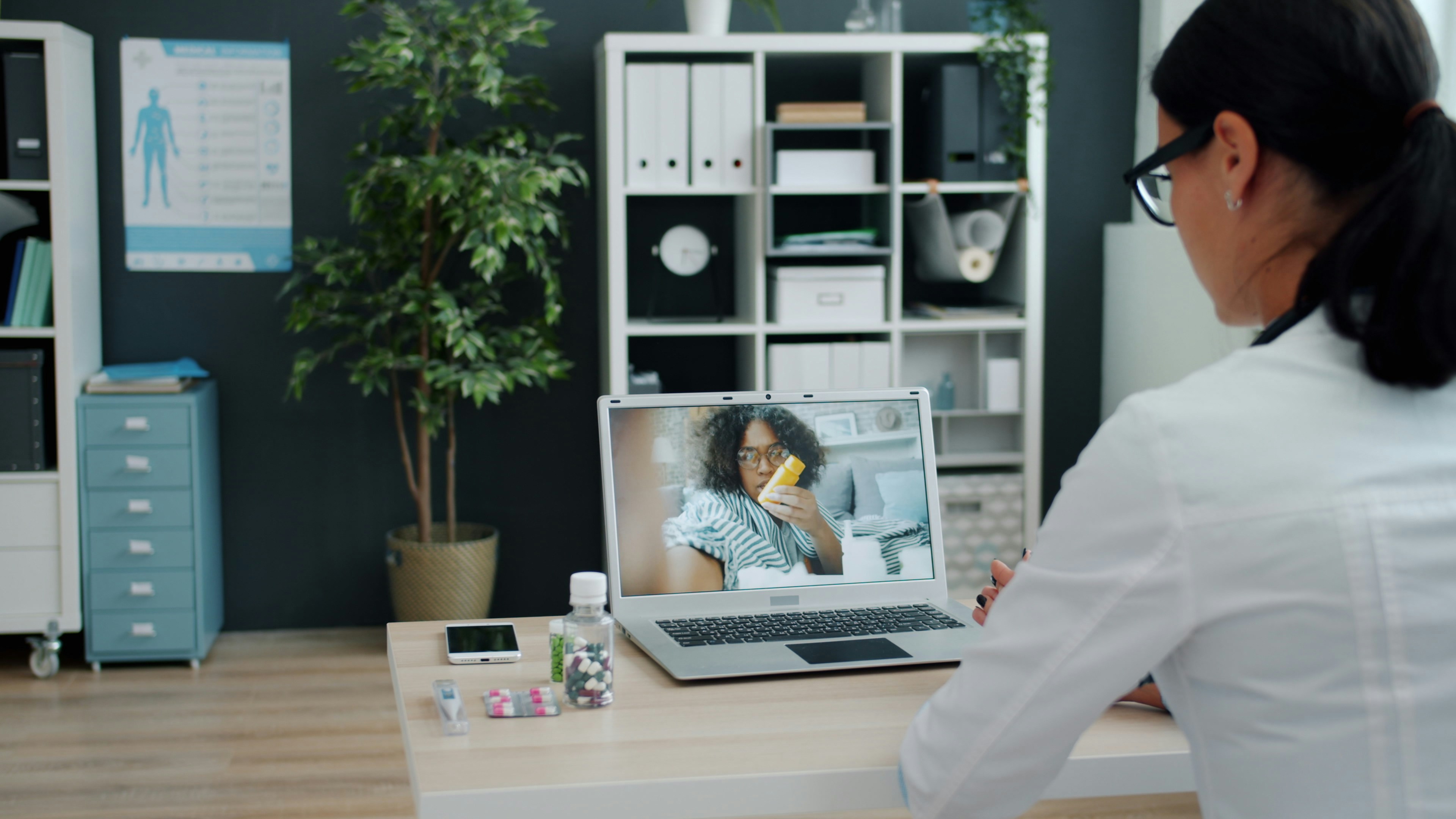 Young woman in doctor's uniform is talking to sick African American girl online using laptop in clinic giving medical advice. People and healthcare concept.