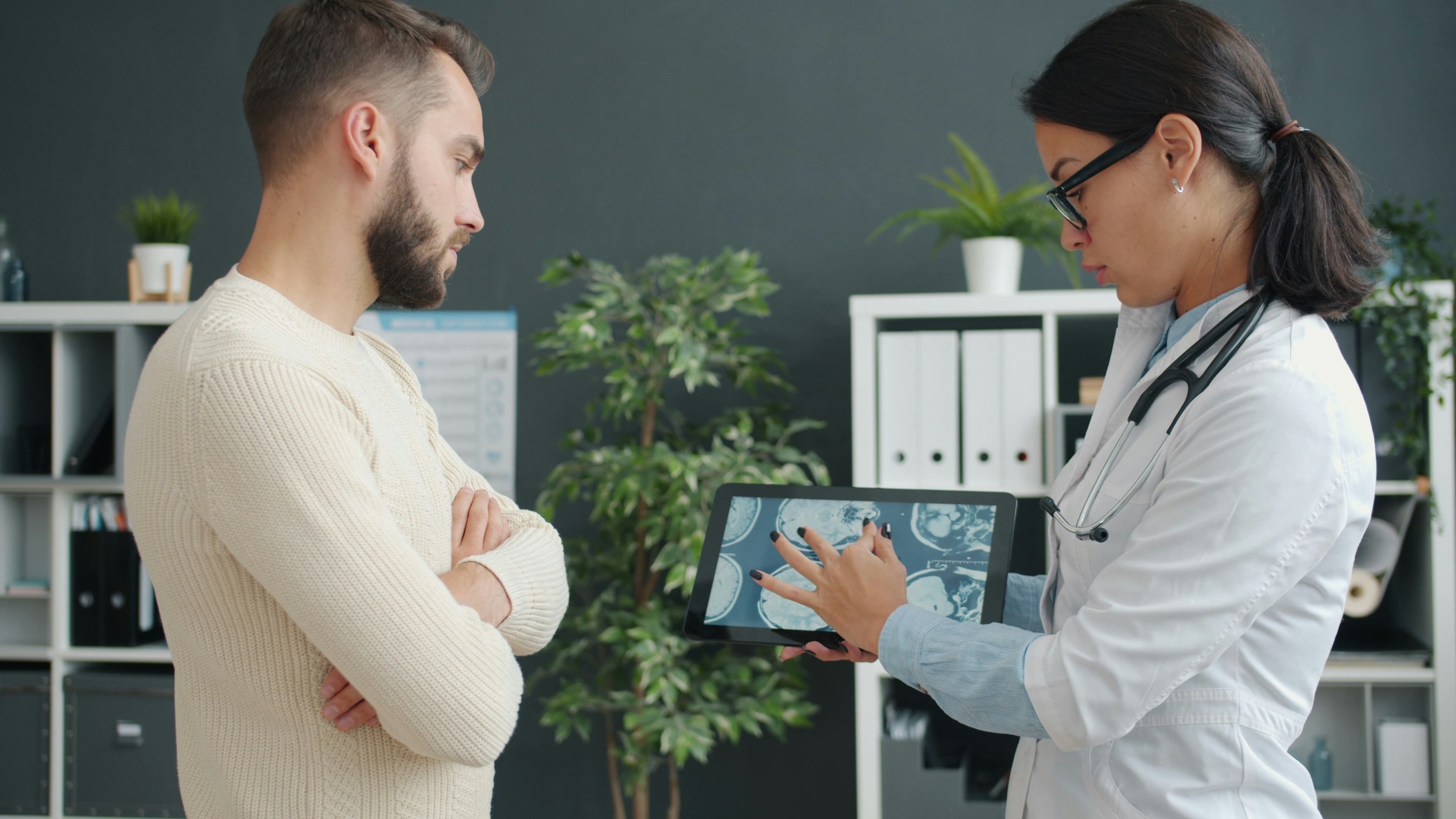 Female practitioner in uniform is showing male patient young man mri results on tablet screen in hospital office. Medical care and people concept.