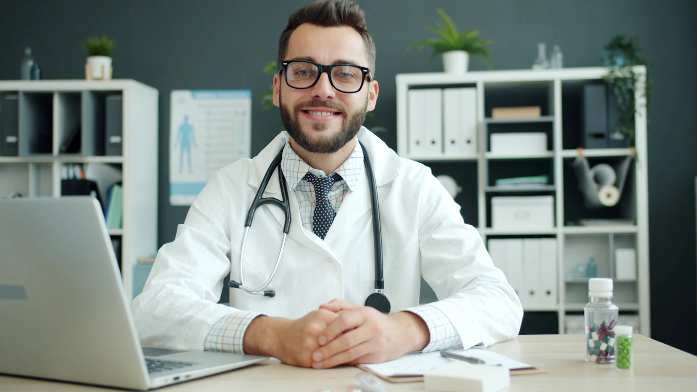 A clinician in a white coat and glasses seated at a desk in a quiet office, the workspace where a private file is reviewed