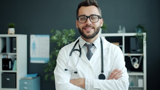 A smiling doctor with glasses and stethoscope stands arms crossed.