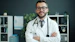 A smiling doctor with glasses and stethoscope stands arms crossed.