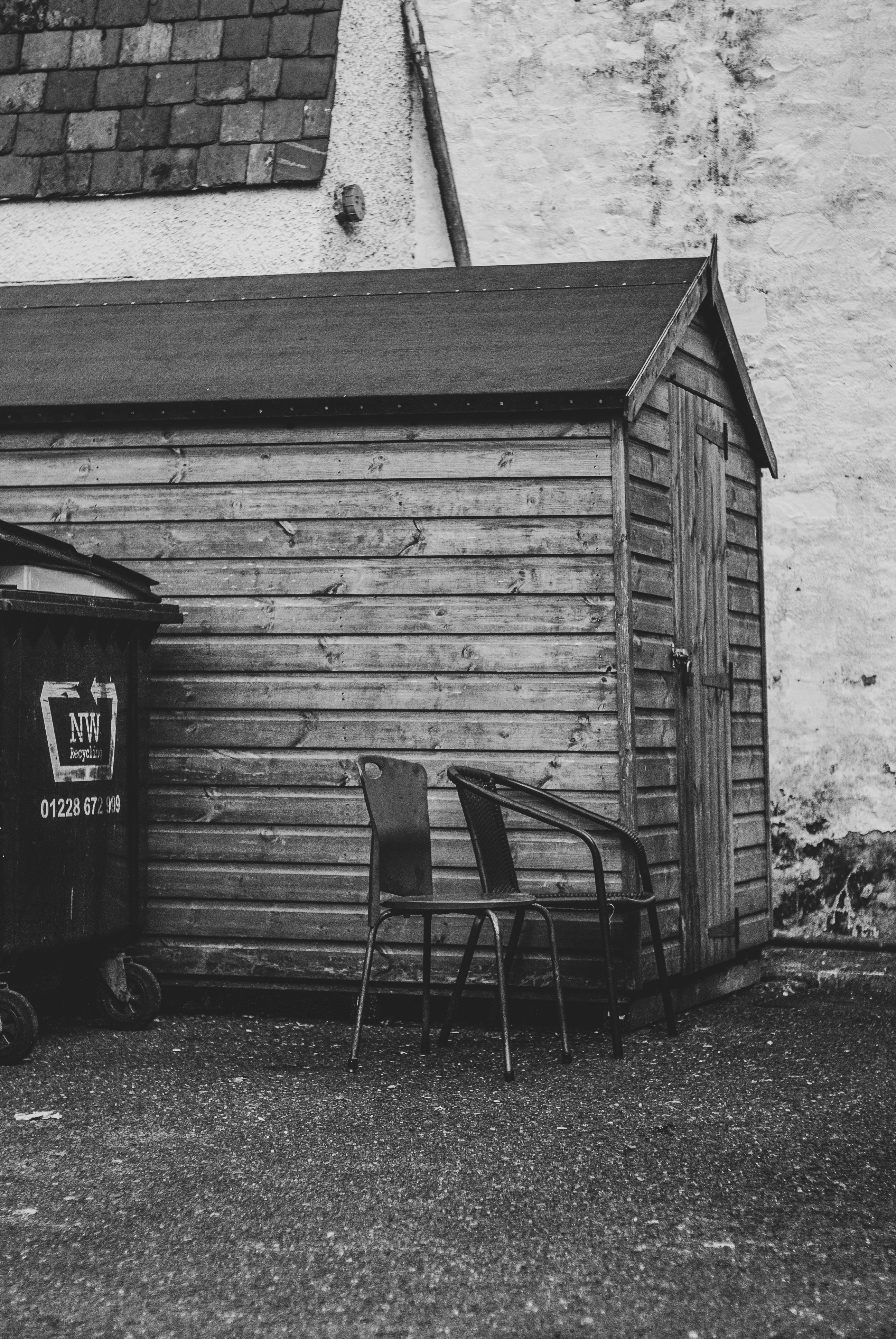 Wooden shed with chairs and trash bin outside.