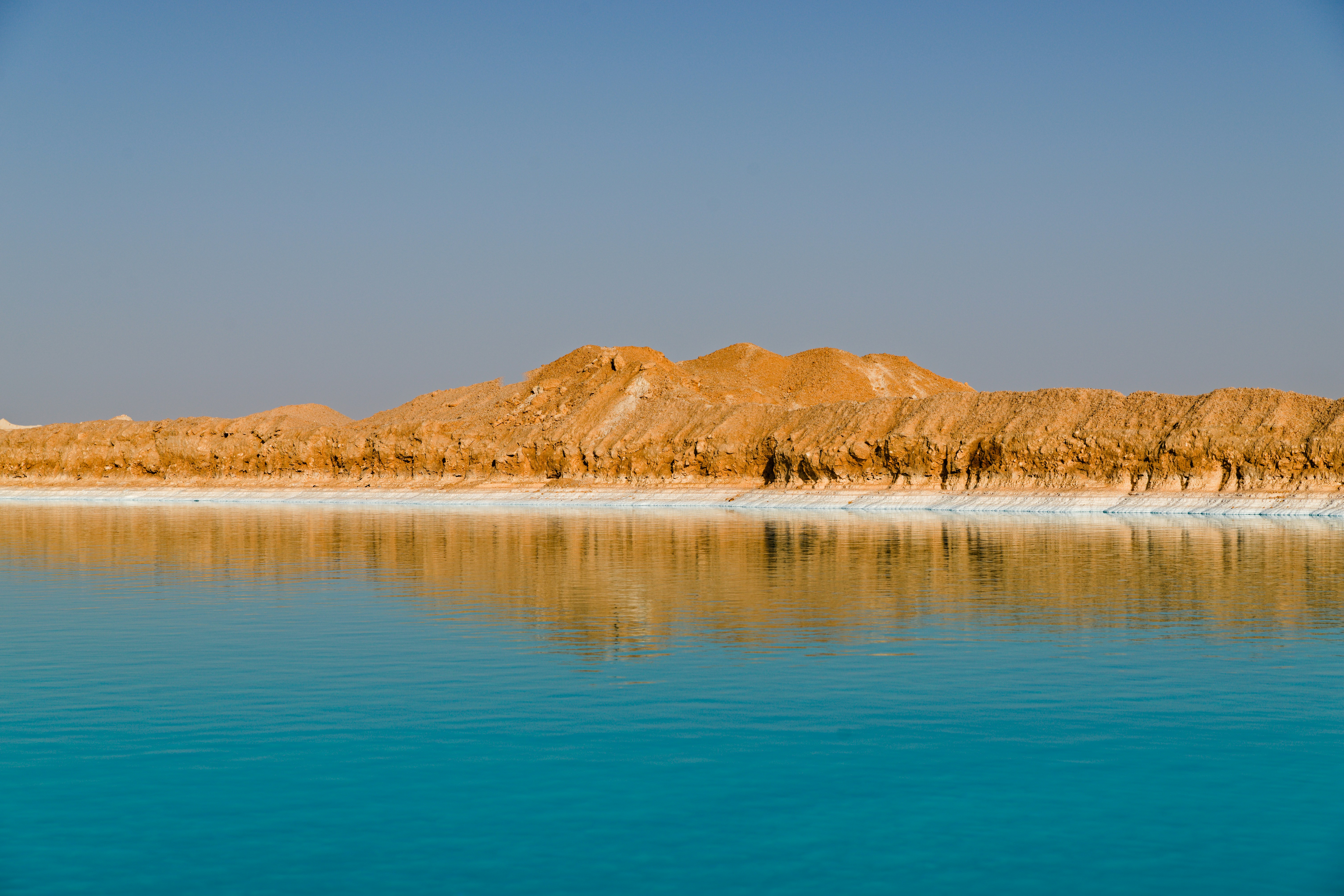 Turquoise water reflects a sandy, rocky shoreline under blue sky.