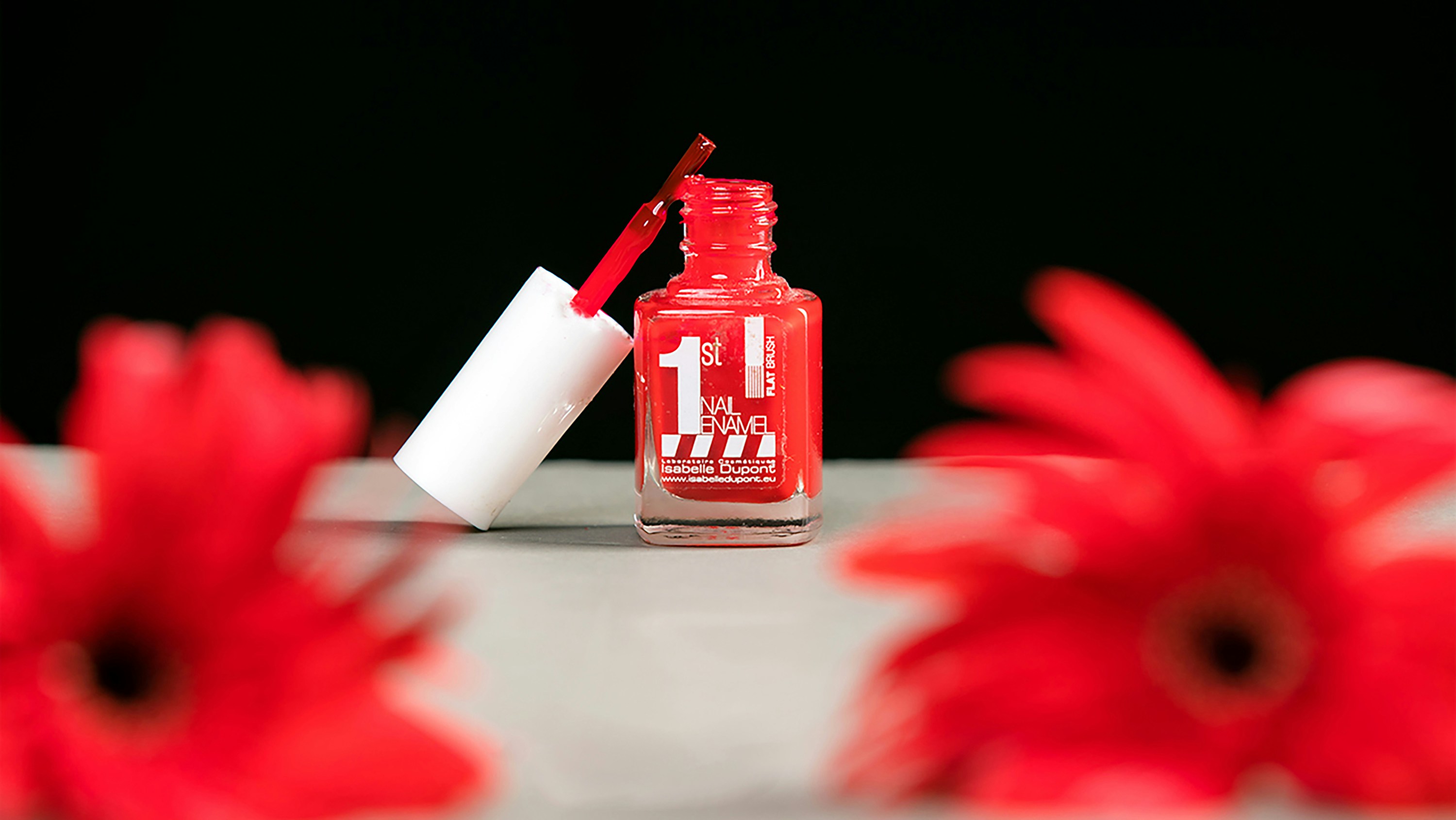 A small, square bottle of red nail polish with its cap and brush resting on the rim. The brush is dripping with polish. In the foreground and background, two red gerbera daisies are out of focus. The background is dark, and the surface is a light gray.