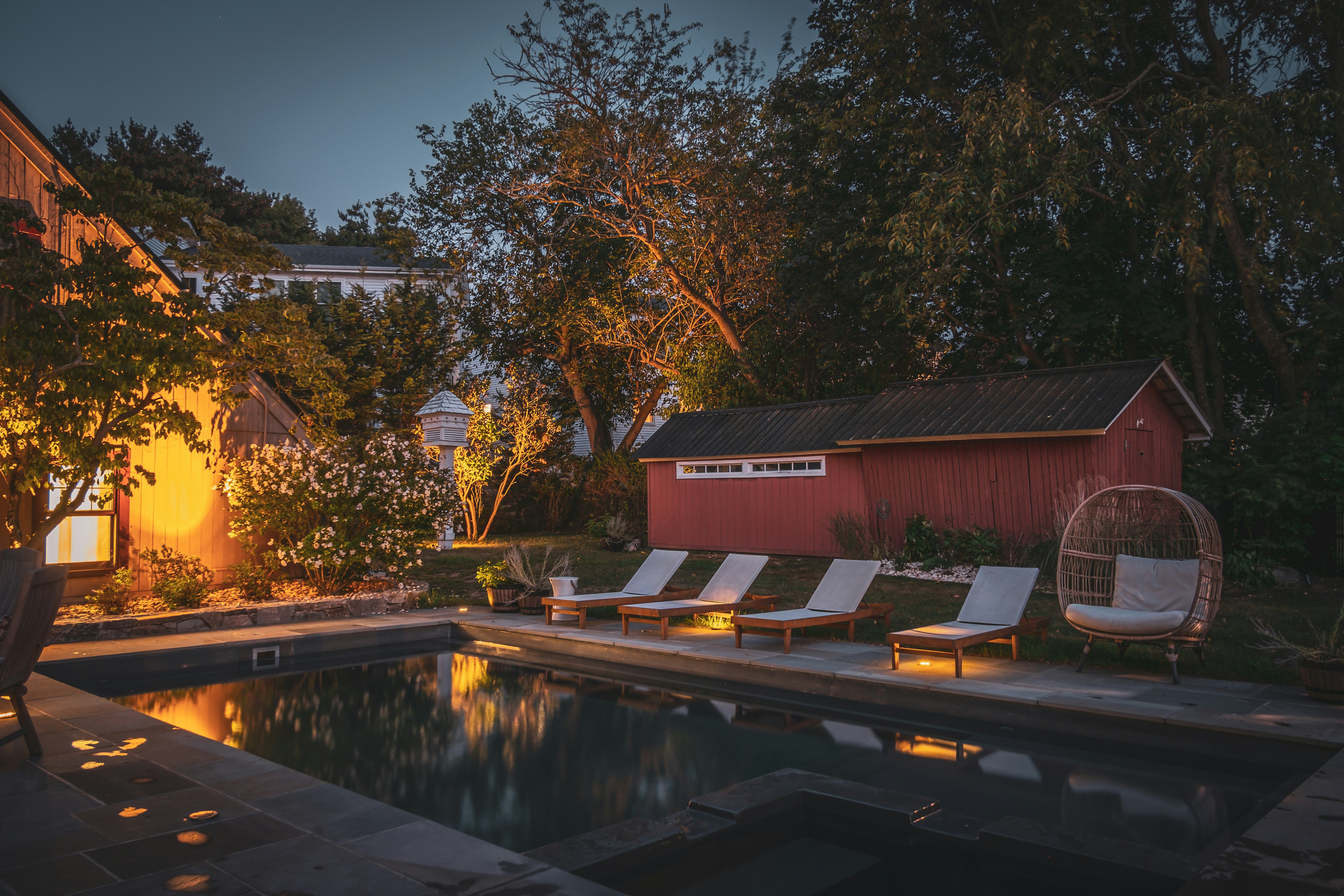 outdoor pool in beautifully decorated backyard surrounded by outdoor white lounge chairs and warm light after sunset