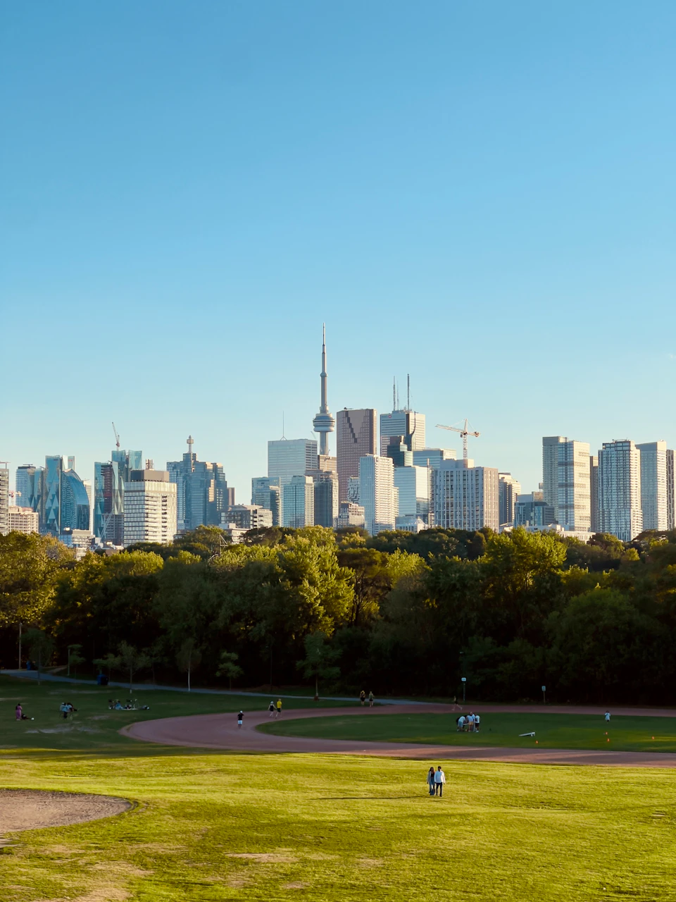 The image depicts a cityscape with a large, modern skyscraper dominating the skyline against a clear blue sky. The buildings are predominantly white and gray, reflecting the urban environment. In the foreground, there is a park filled with lush green trees that provide a natural contrast to the man-made structures. The park is populated by people enjoying various activities such as walking or sitting on benches. A winding path runs through the center of the park, adding an element of movement and dynamism to the scene.

The image is taken from a high vantage point, offering a panoramic view of the city's skyline and the park below. The sun shines brightly in the sky, casting shadows that add depth and dimension to the scene.