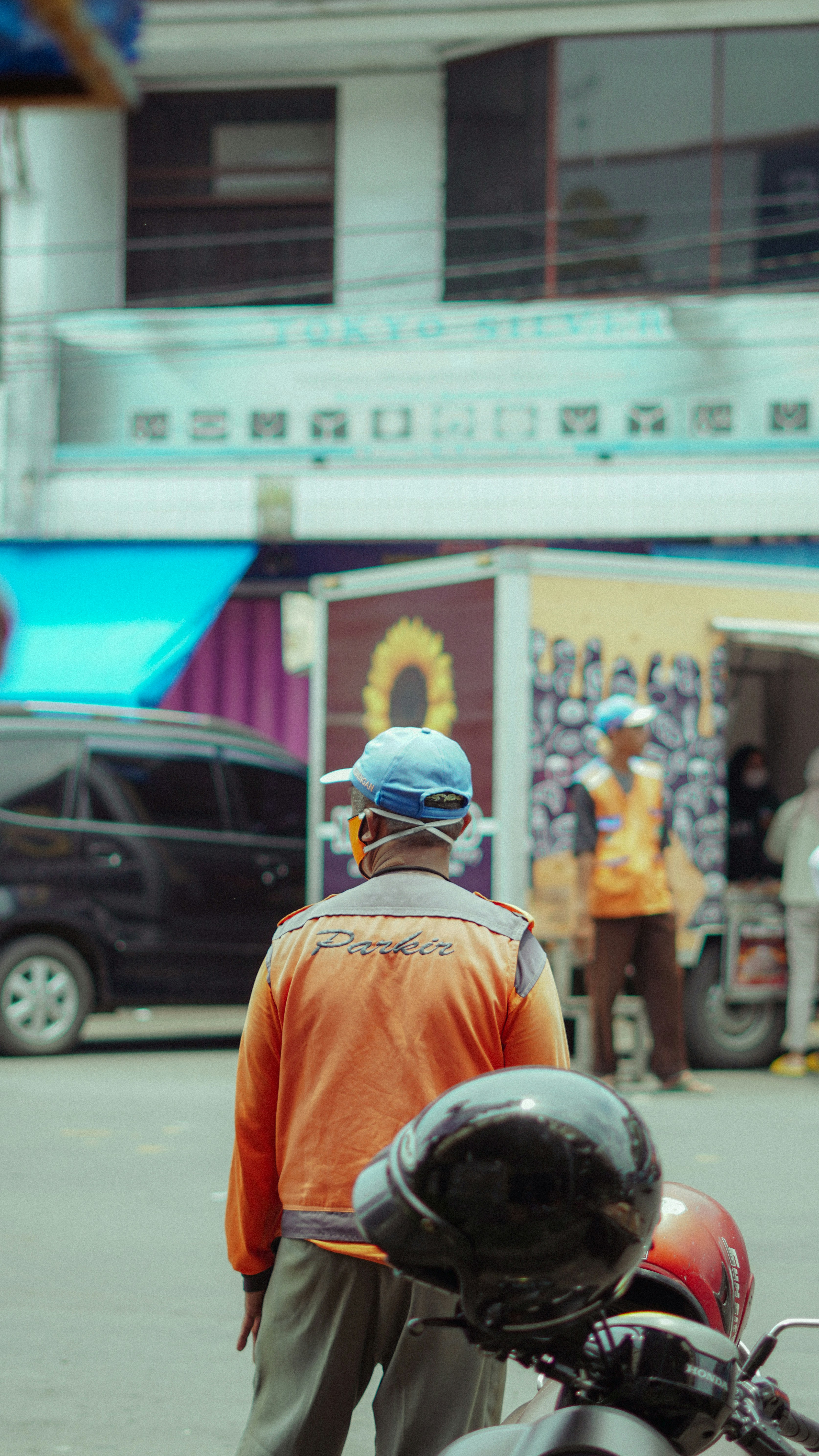 Man in orange jacket and blue cap stands near motorcycle.
