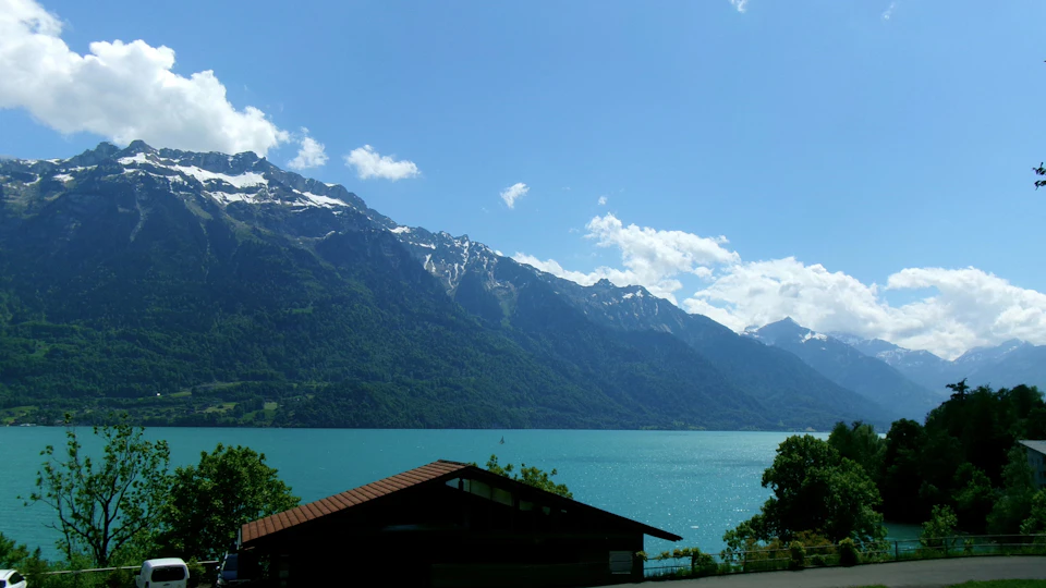 The image depicts a serene mountain landscape with a large lake nestled amidst the mountains, surrounded by lush green trees and shrubs. The sky above is a clear blue, dotted with fluffy white clouds that add to the picturesque scene. In the foreground, there are several buildings, including a small wooden cabin with a brown roof, situated on the right side of the image.