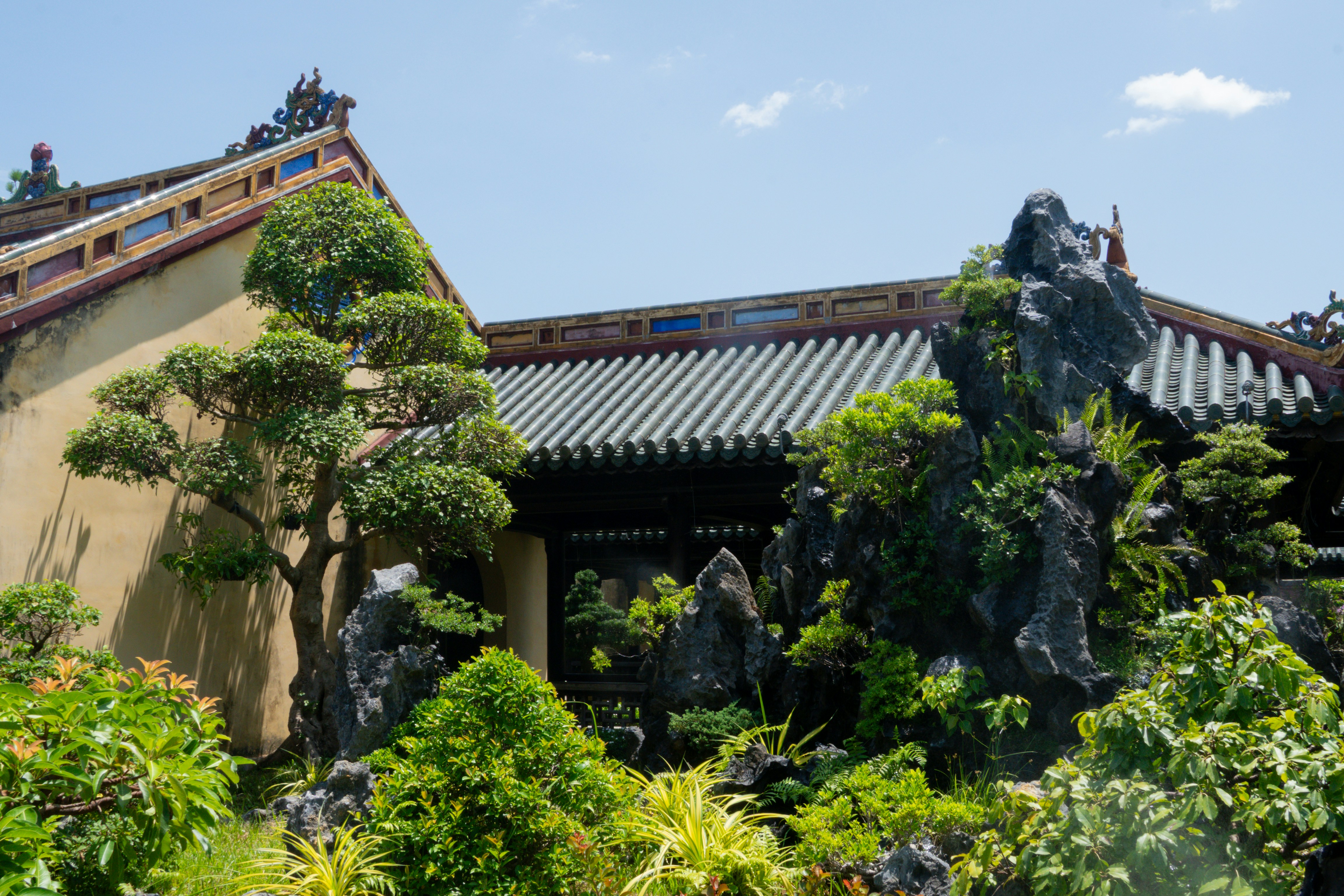 A traditional Asian garden featuring a building with a grey tiled roof and yellow walls, surrounded by lush green vegetation, bonsai trees and large rock formations. 