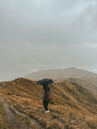 A hiker in a dark vest holds a black umbrella on a grassy ridge path under low grey cloud, bare autumn hills stretching away