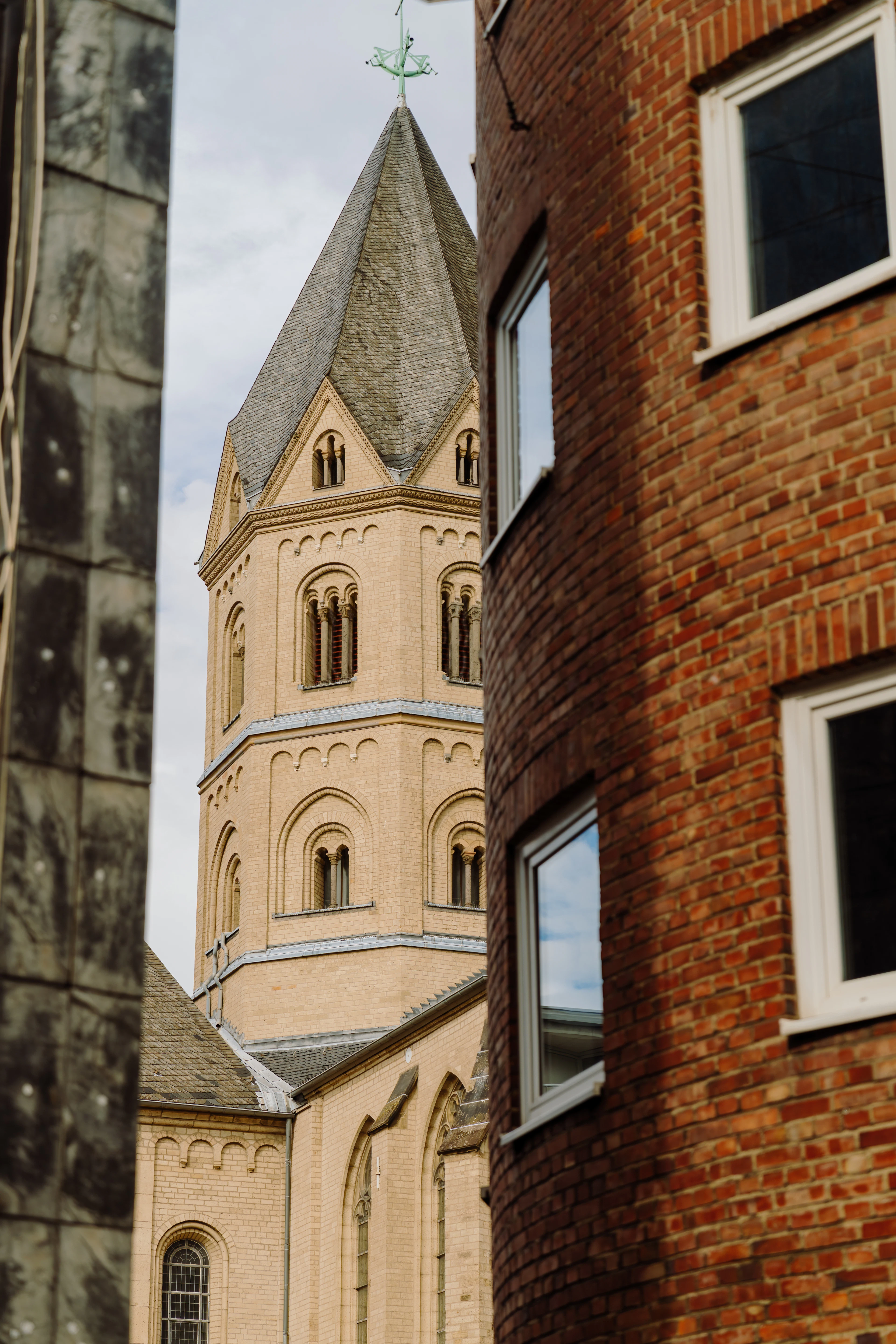A Romanesque church tower with a steep pointed spire glimpsed between two buildings in Cologne's old town.