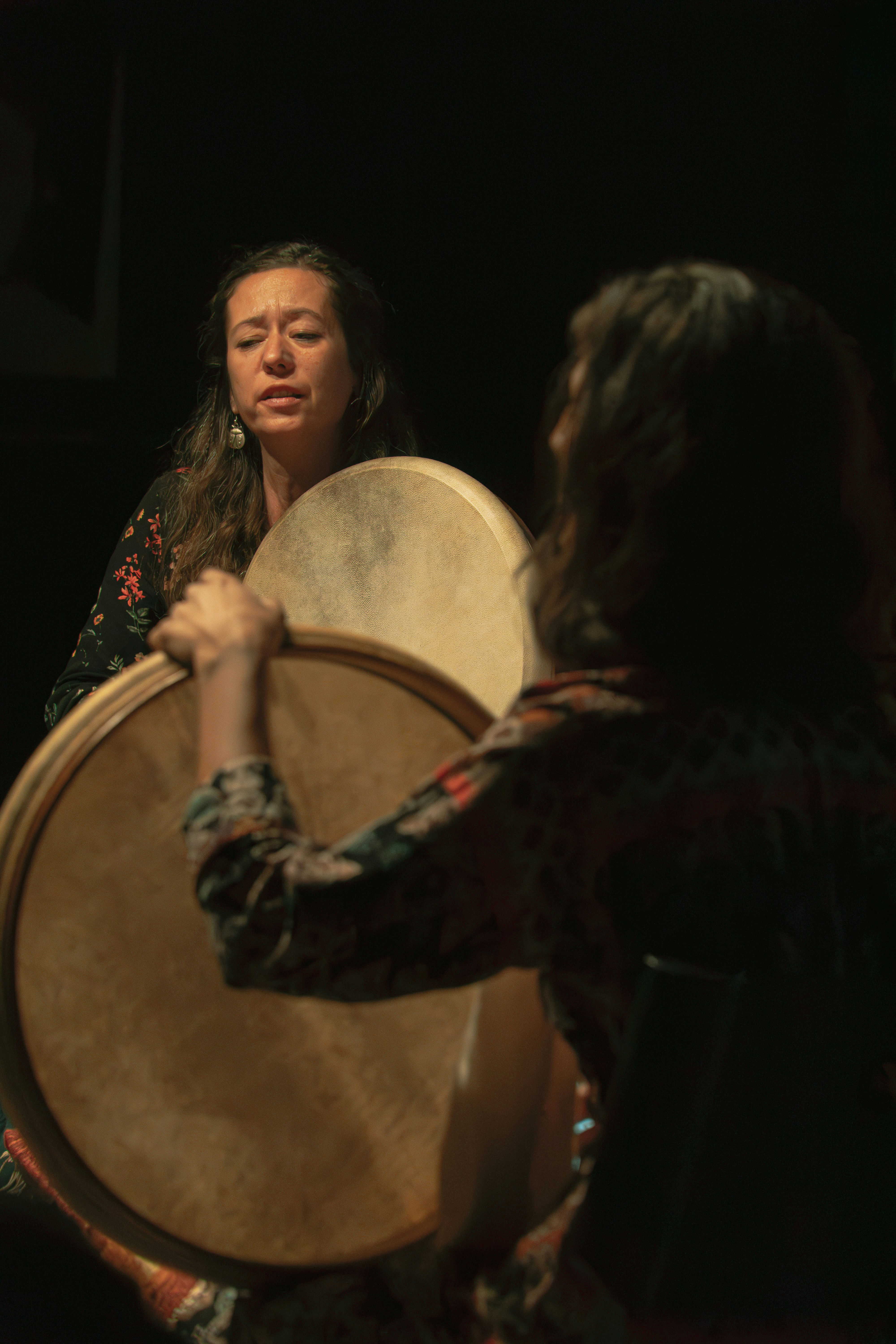 Two women playing large round frame drums in a dimly lit room