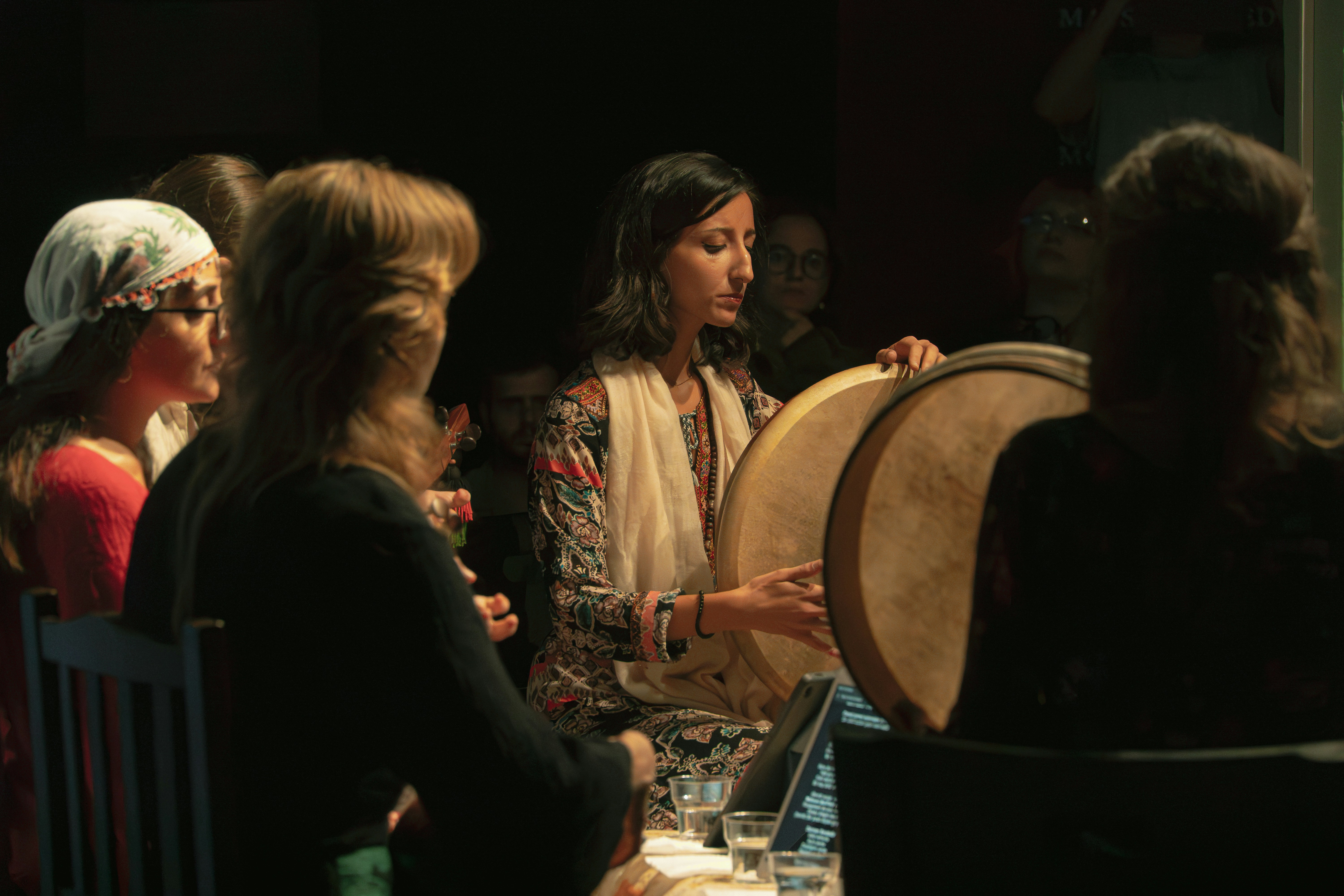 A woman playing a frame drum in a dimly lit room with other musicians