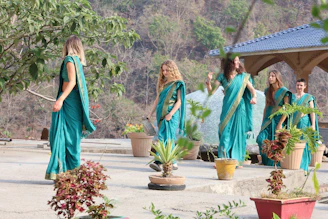 Women in teal saris dancing outdoors near potted plants.
