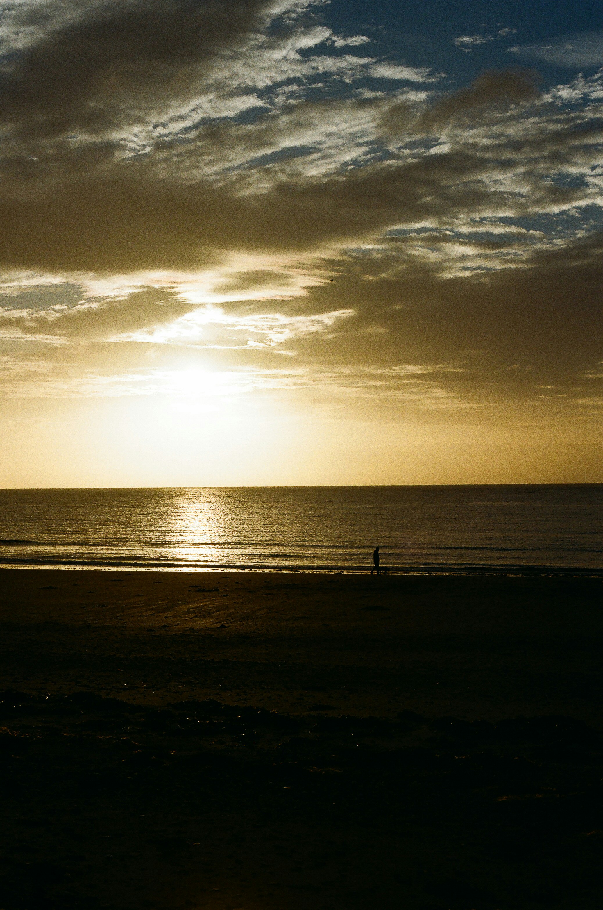 Lone figure walks on beach at sunset with dramatic clouds.