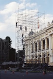 Workers climb scaffolding around a building facade.