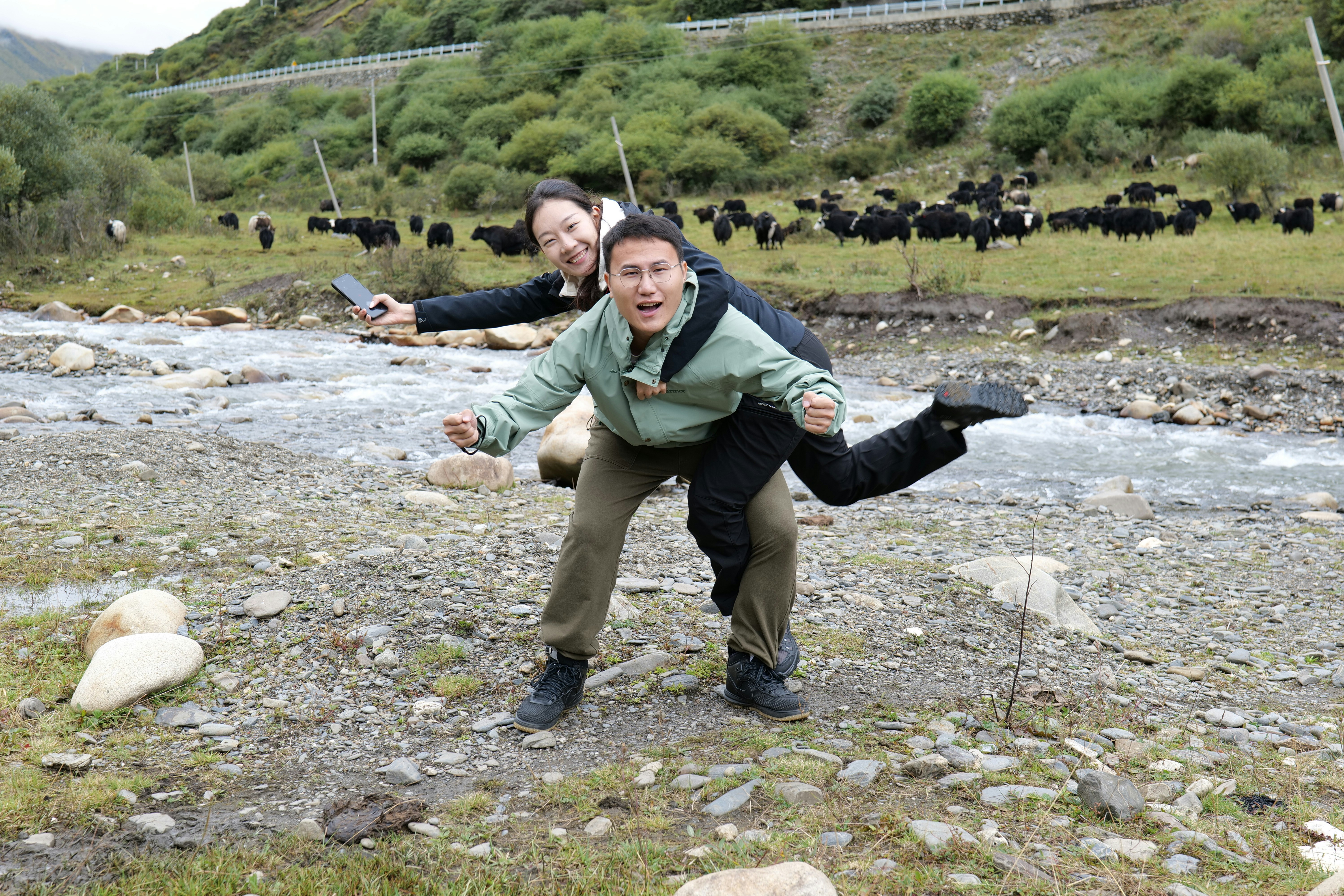 Two men piggyback riding near a stream with yaks.