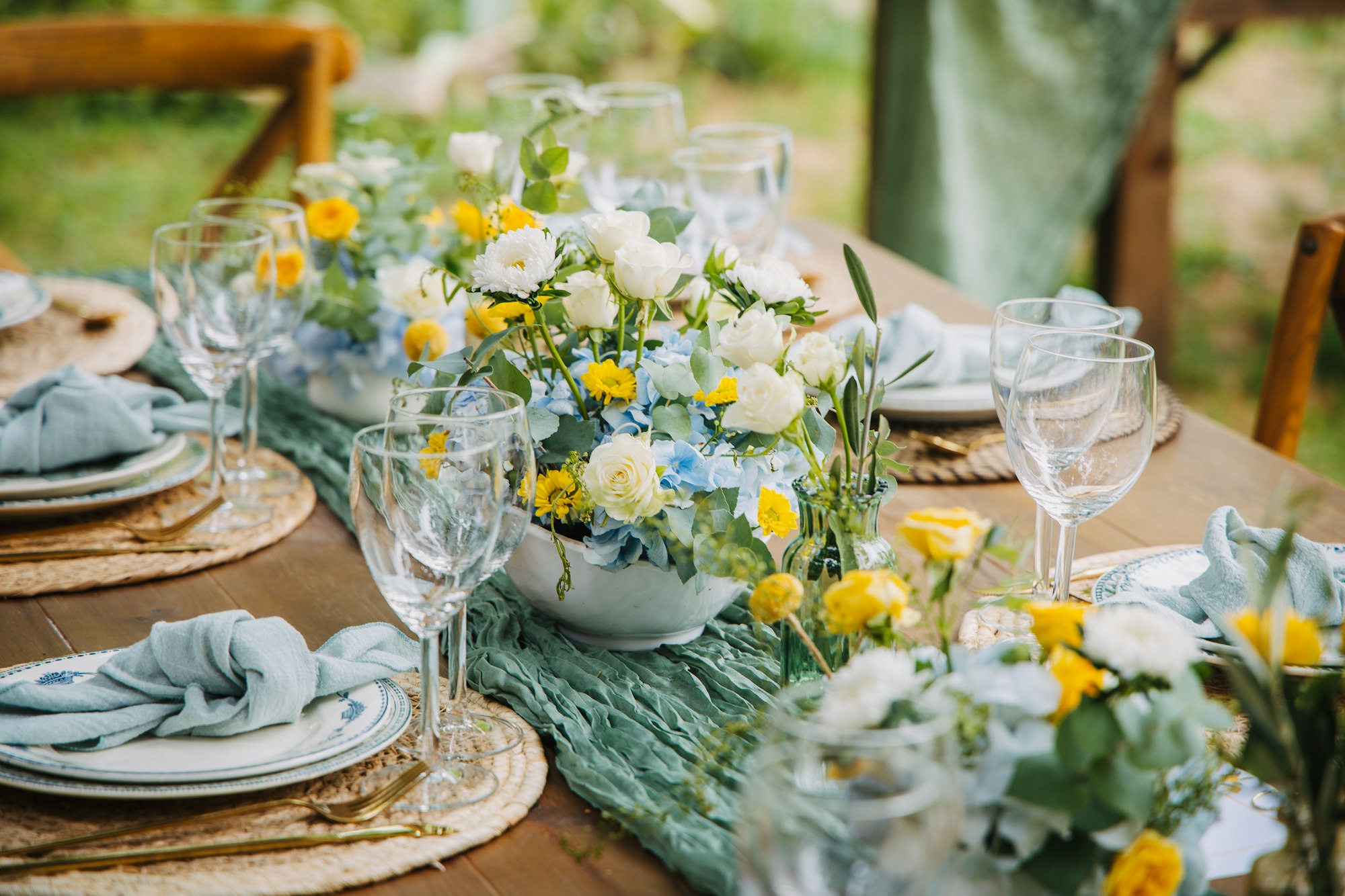 A long outdoor dining table beautifully set with floral centerpieces and greenery arrangements under soft natural light