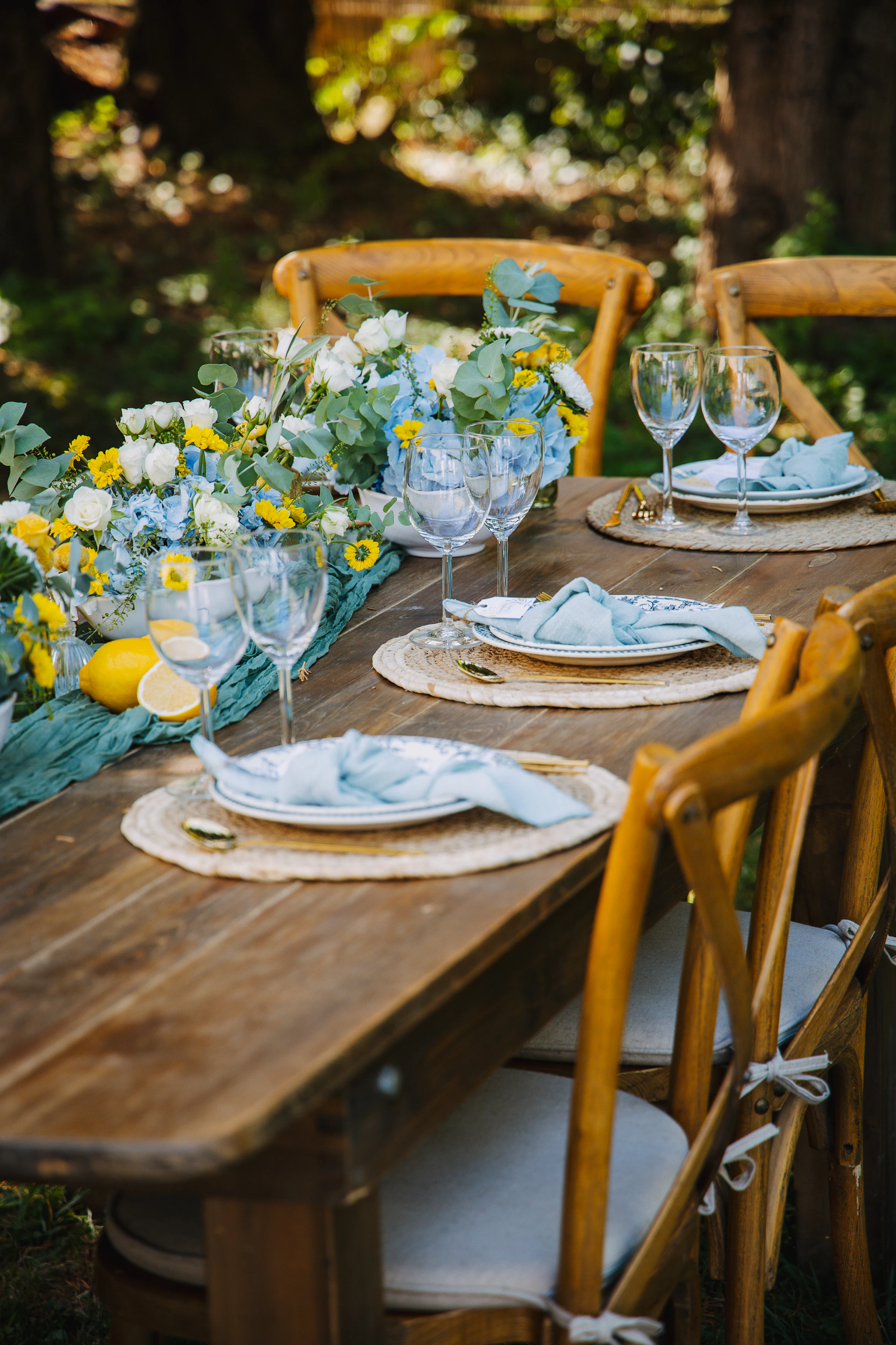 Long outdoor table prepared with plates, cloth napkins, glasses, flowers, and lemons