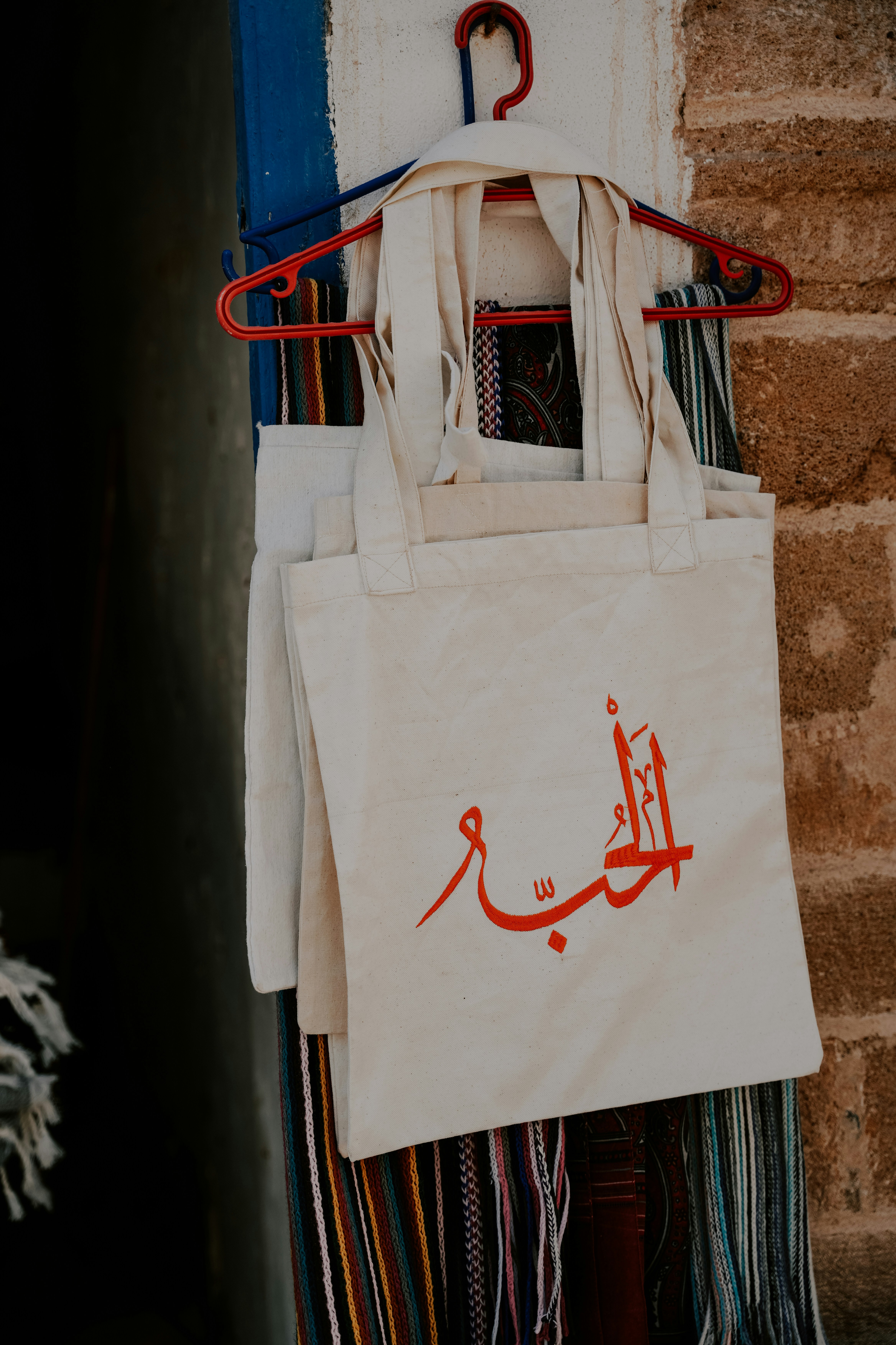 White tote bag with red arabic script hangs on hanger.