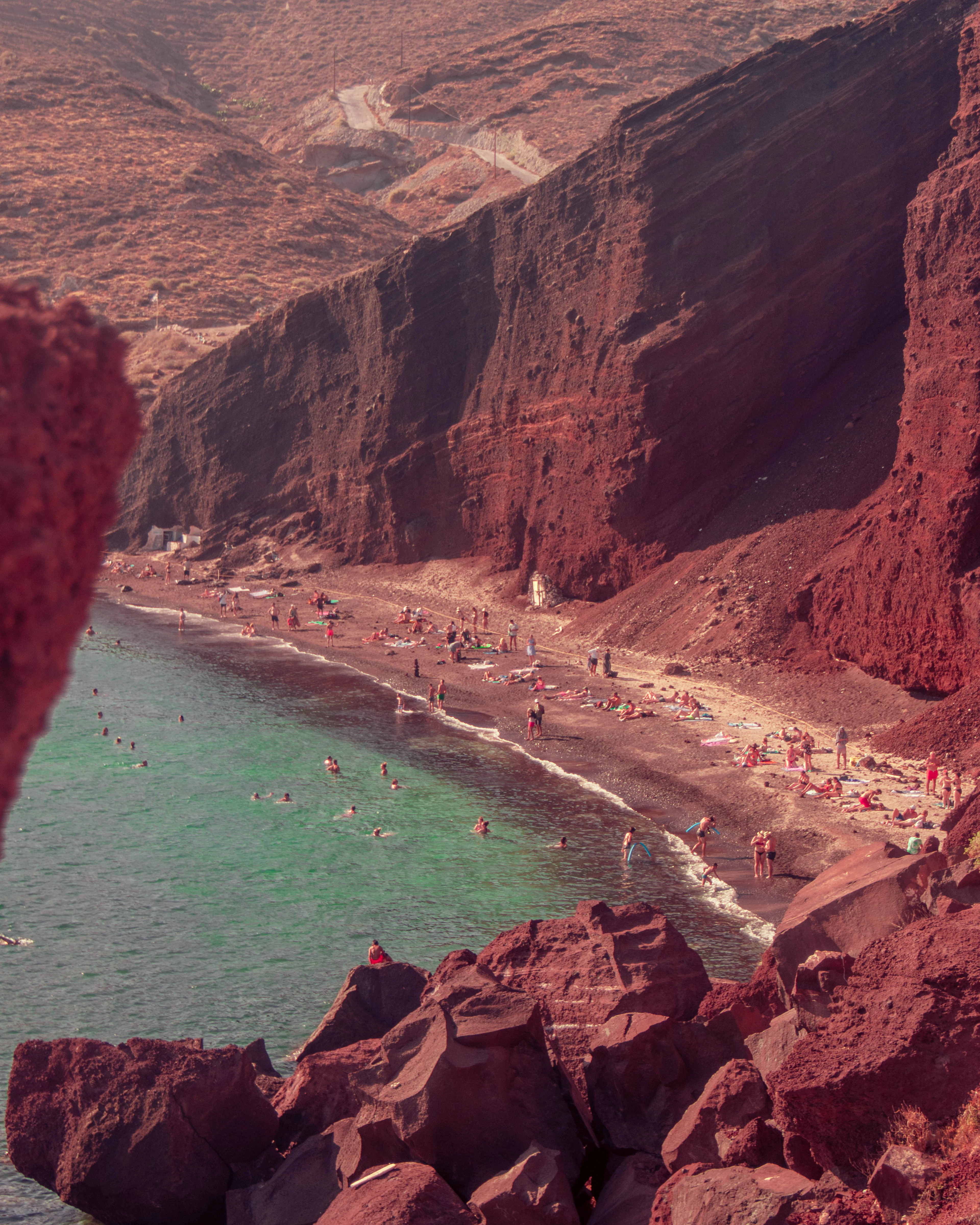 Bathers on the dark volcanic shingle of Santorini's Red Beach, enclosed by towering rust-red lava cliffs above clear turquoise water