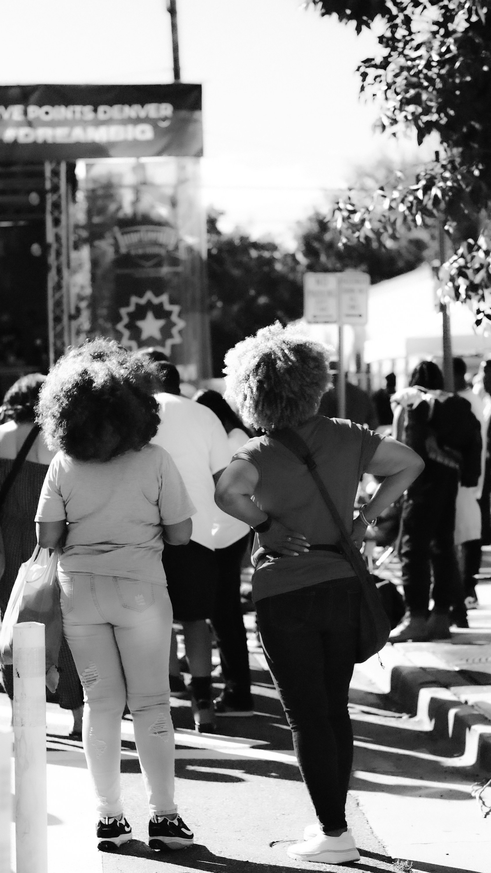 Two women stand on a street with people behind them.