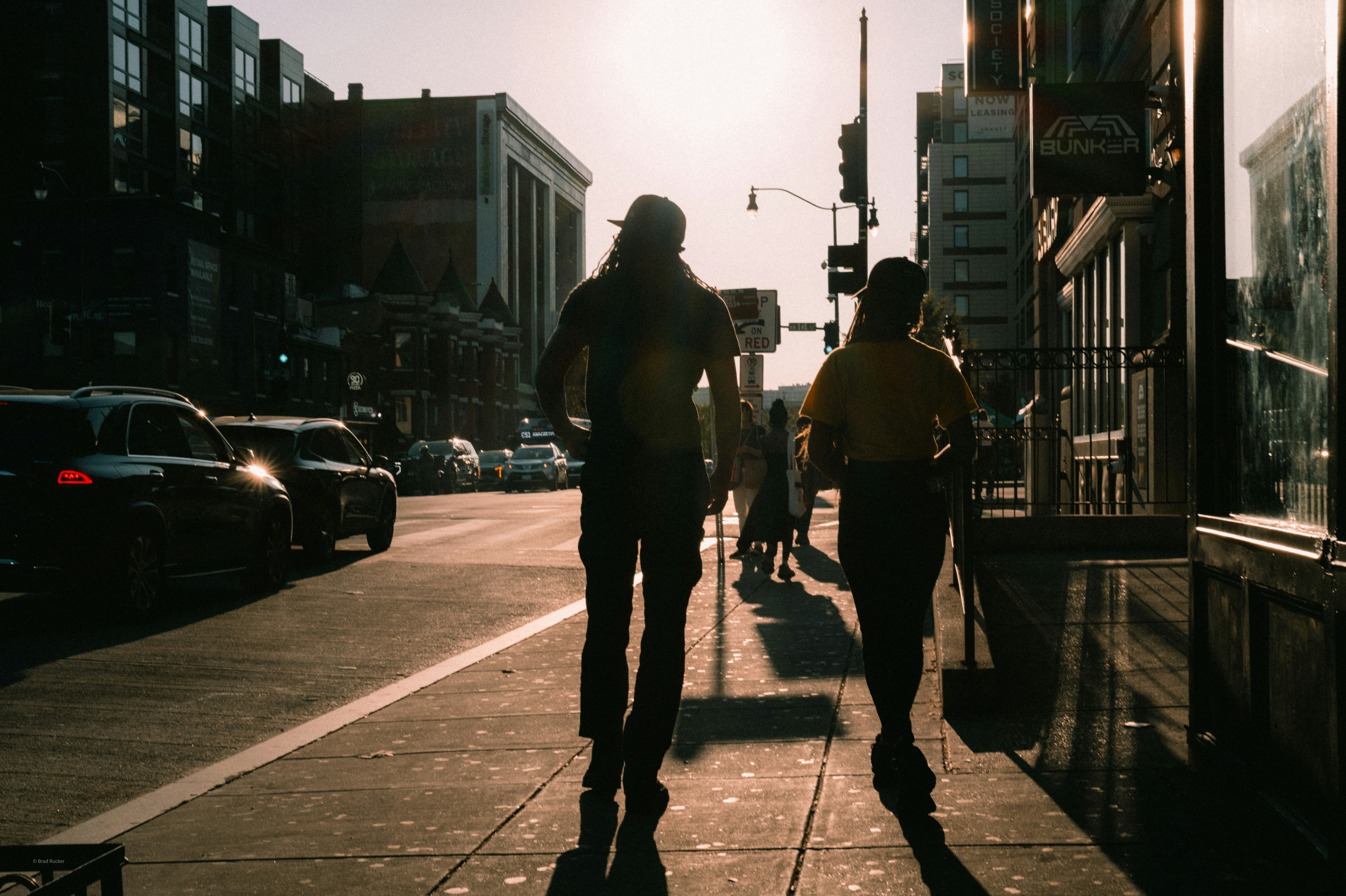 Two people walk down a sunlit city sidewalk.
