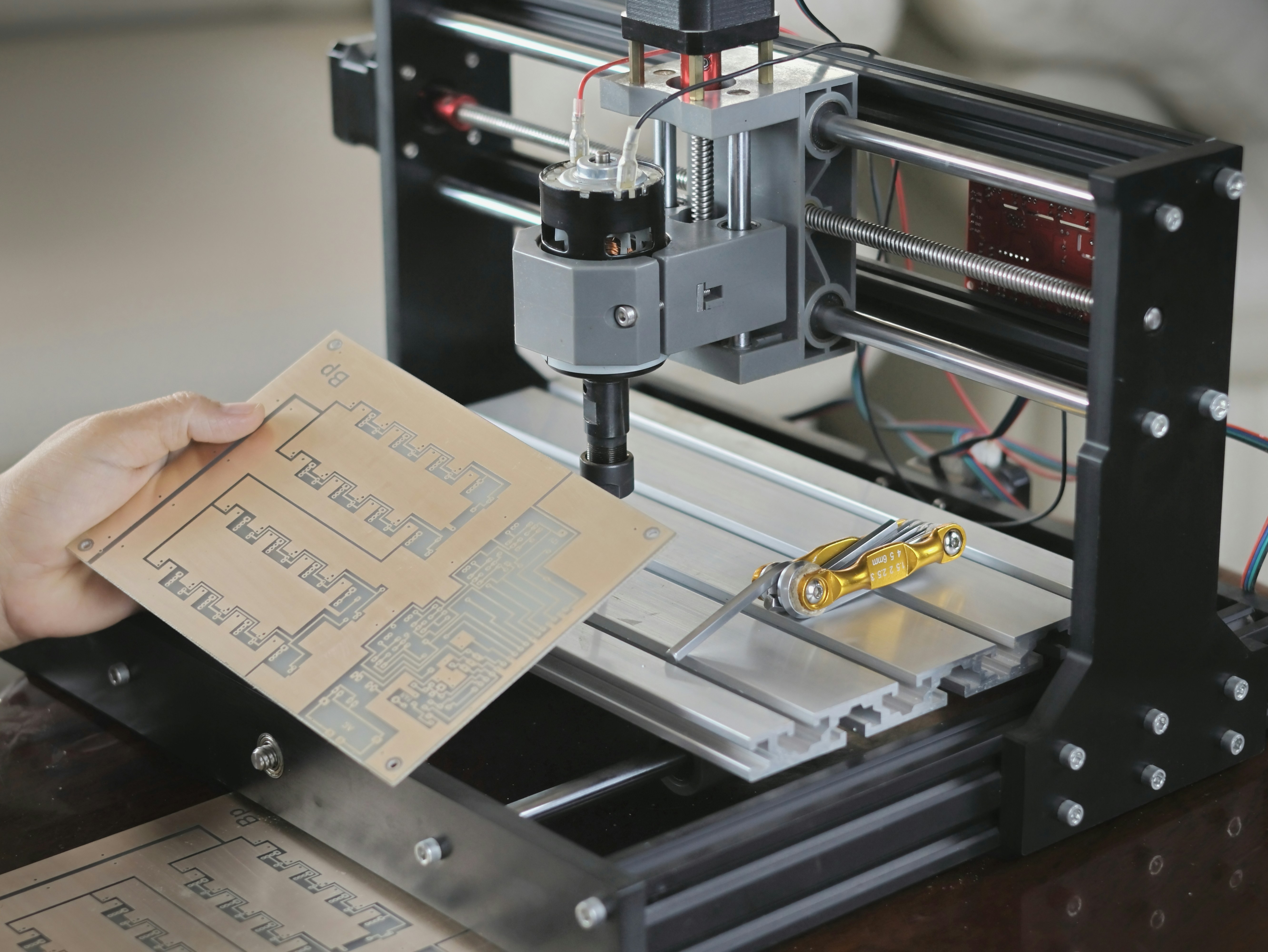A close-up of a printed circuit board (PCB) being prepared for engraving by a small CNC machine in operation. This demonstrates modern manufacturing technology and precision, with a hand holding the completed circuit board. Suitable for media related to electronics