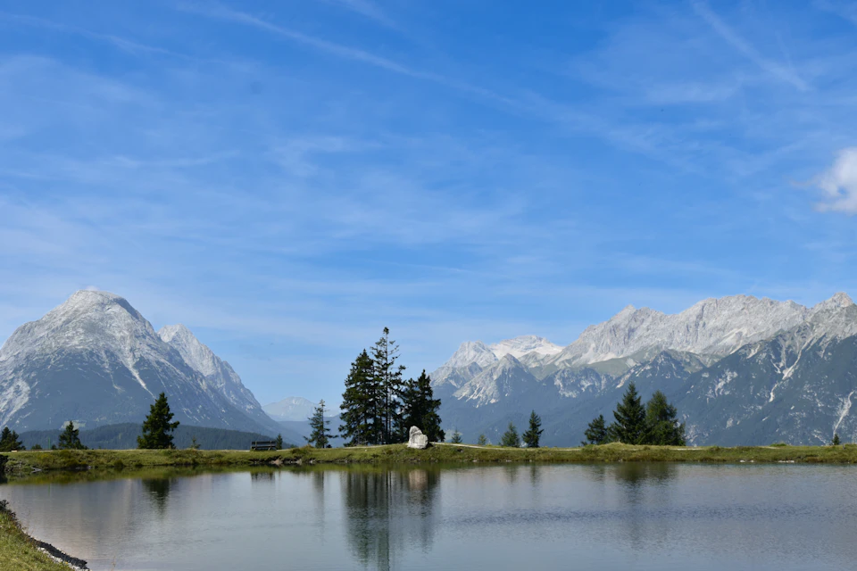 The image depicts a serene mountain landscape with a large lake at its center, surrounded by snow-capped mountains and clear blue skies. The mountains are covered in white snow, creating a stunning contrast against the sky. In the foreground, there is a small island of trees and bushes, adding a touch of green to the otherwise monochrome scene. The image is taken from a high vantage point, providing a comprehensive view of the landscape below.