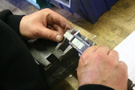 DYEBACK Hands using a digital caliper to measure metal part.