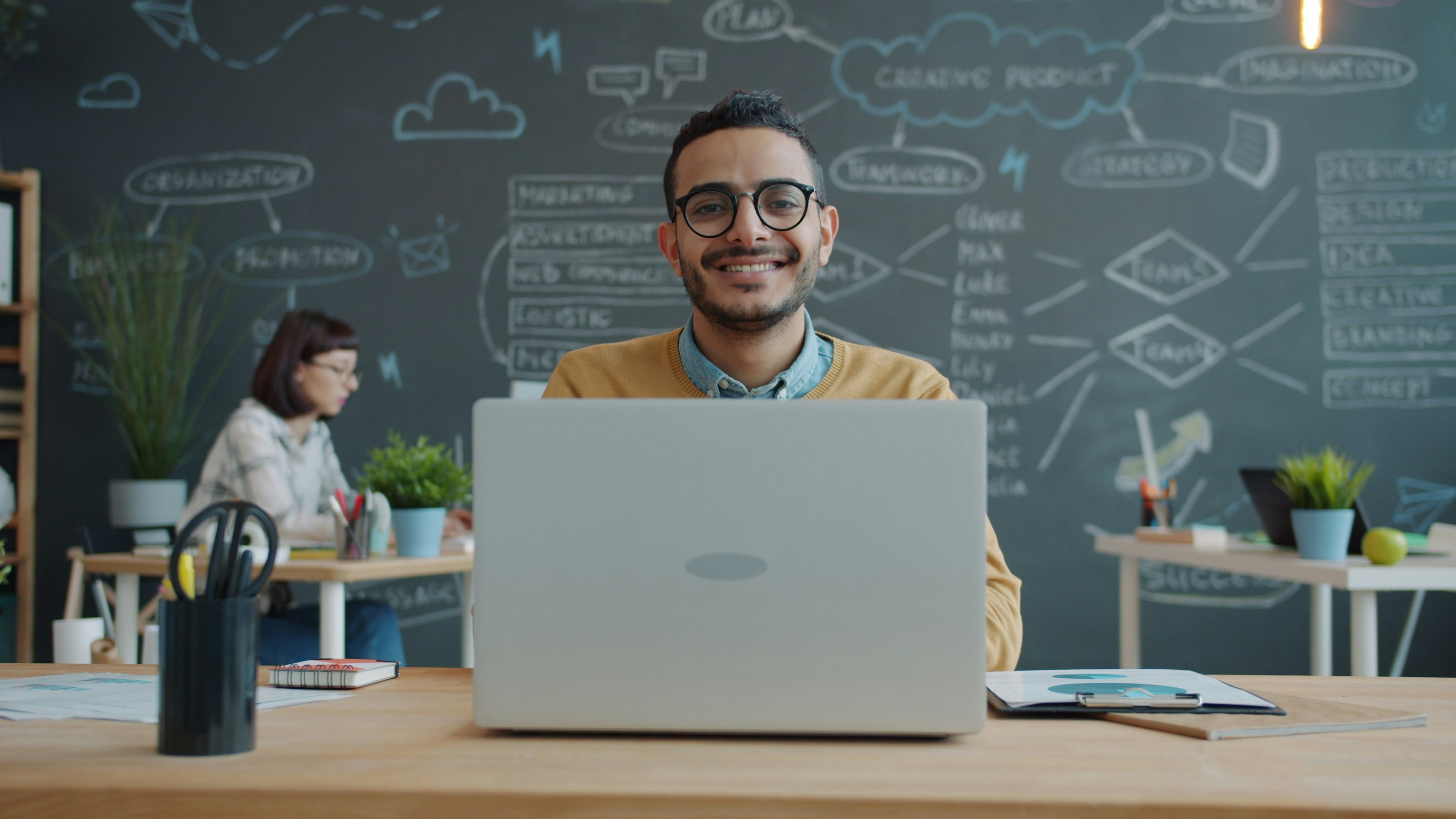 Portrait of happy Middle Eastern man sitting at desk with laptop in office and smiling looking at camera. People, job and modern technology concept.
