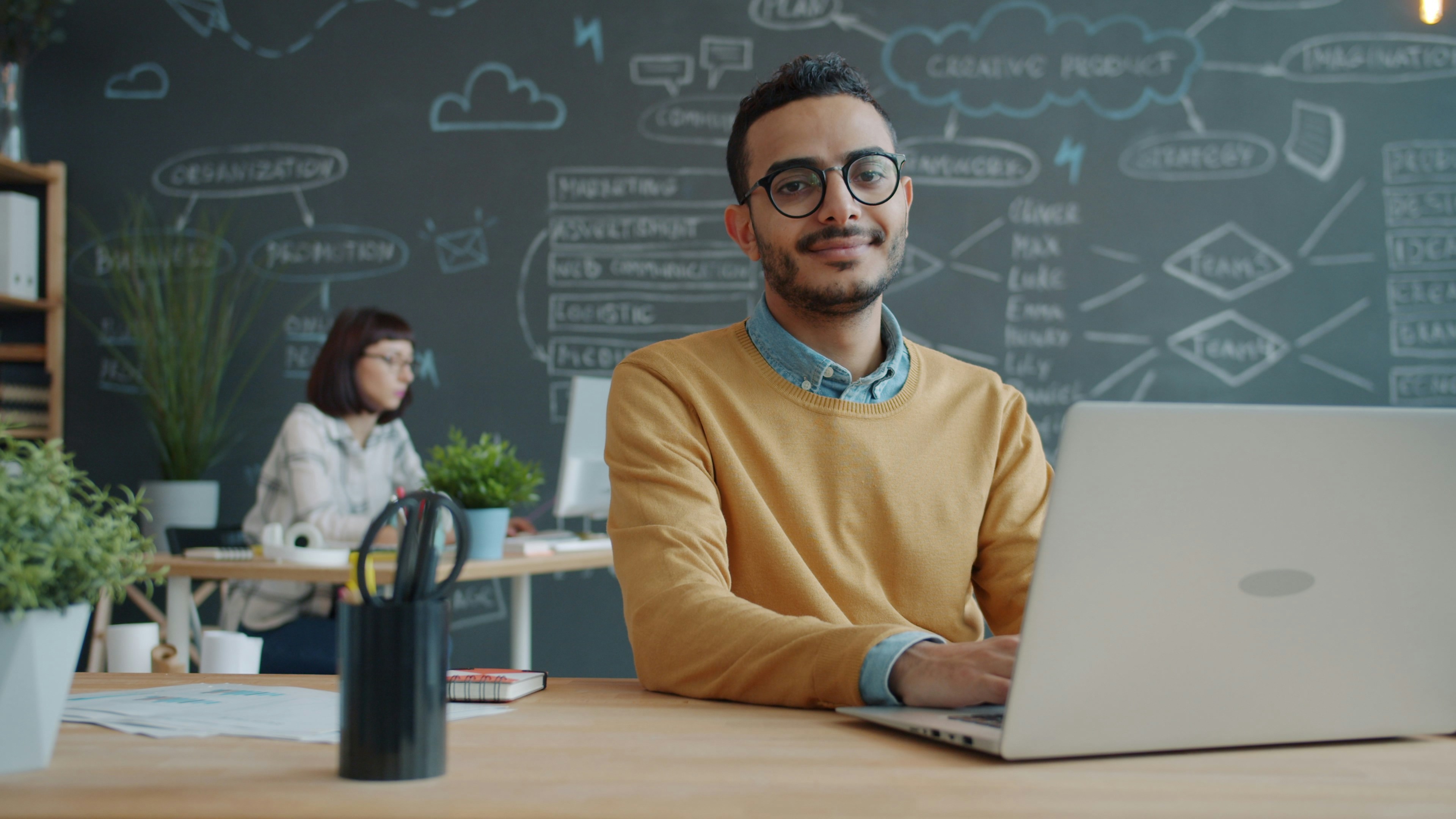 Portrait of cheerful Middle Eastern man at desk with laptop in modern office sitting alone and looking at camera smiling. People, work and emotions concept.