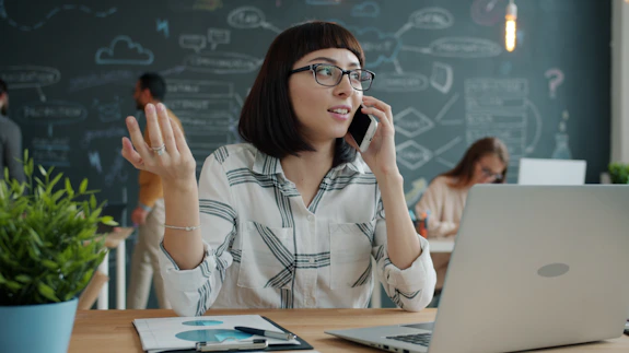 Woman talking on phone at desk with laptop and plant.