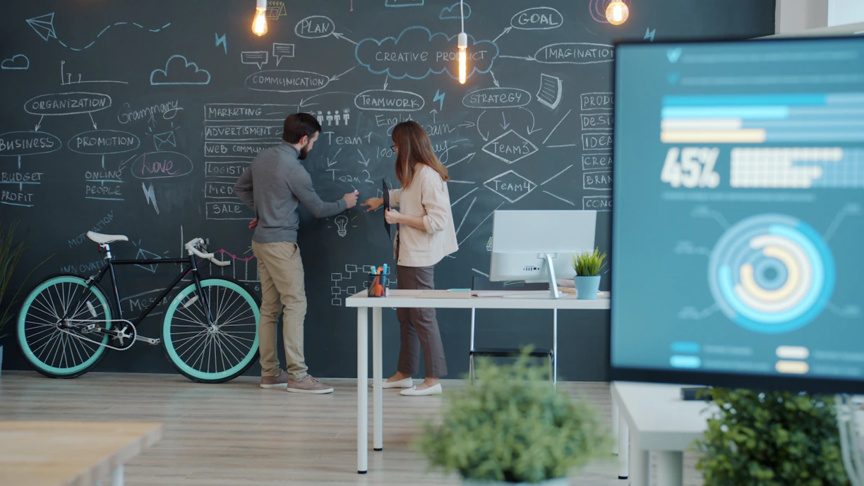 Two digital marketers collaborating on a chalkboard wall with a bicycle leaning up against the wall.