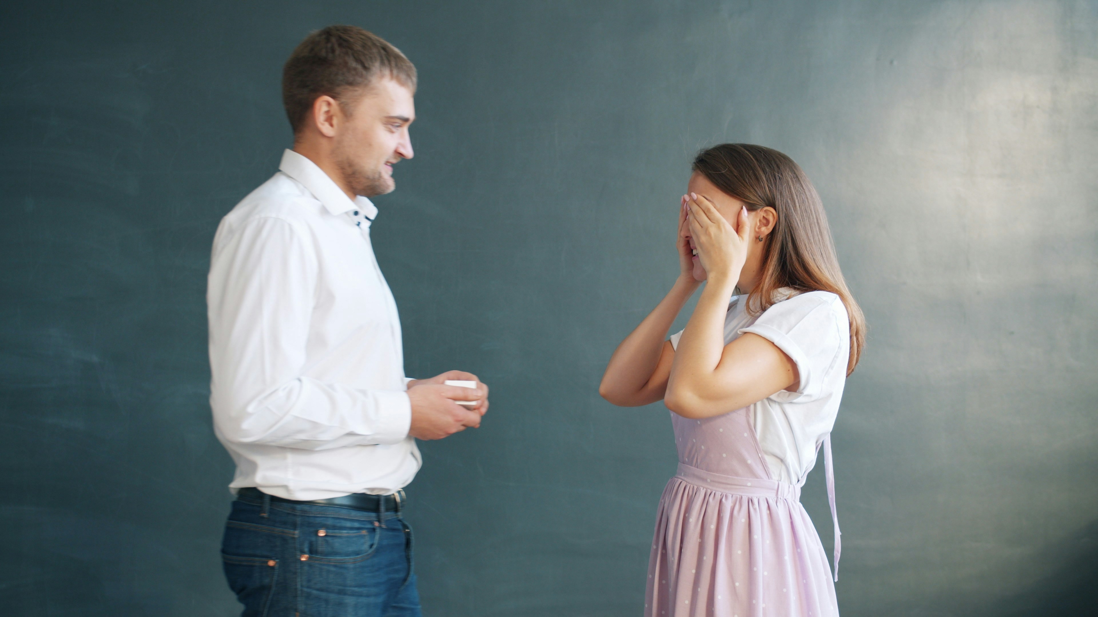 Good-looking man is proposing to pretty girl giving engagement ring in jewellery box, young lady is excited. Couple is standing against gray background.