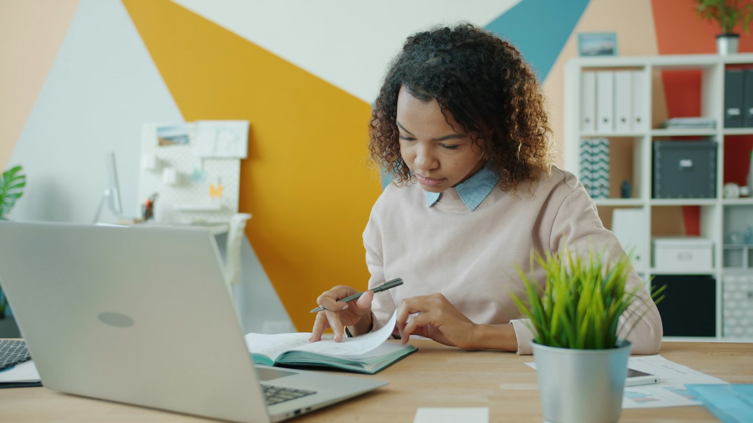 A woman at an office desk writes in a notebook beside her open laptop, studying and planning her next career step