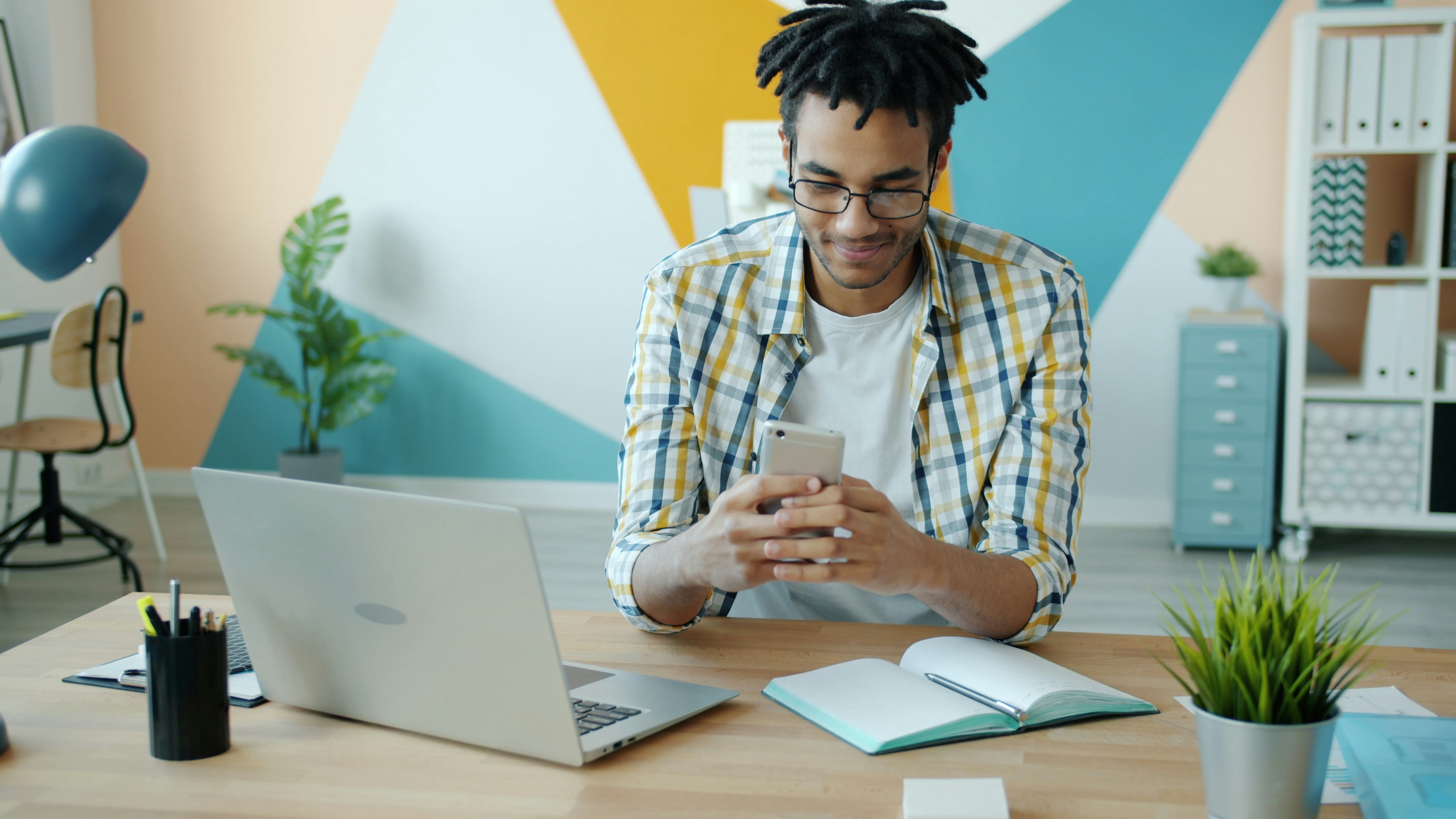 Man looking at phone at desk with laptop and plants.