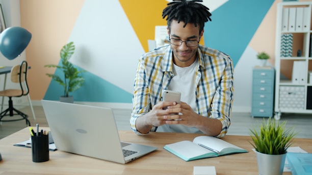 Man looking at phone at desk with laptop and plants.