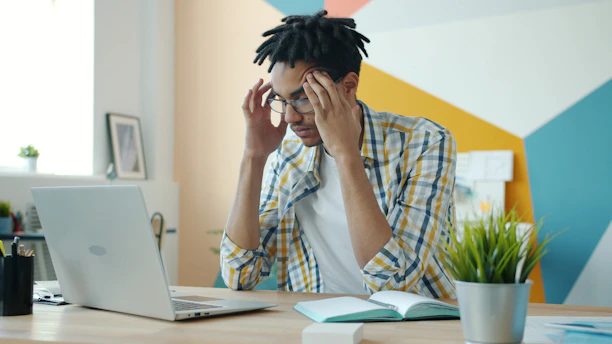 Man with dreadlocks holding head at desk with laptop