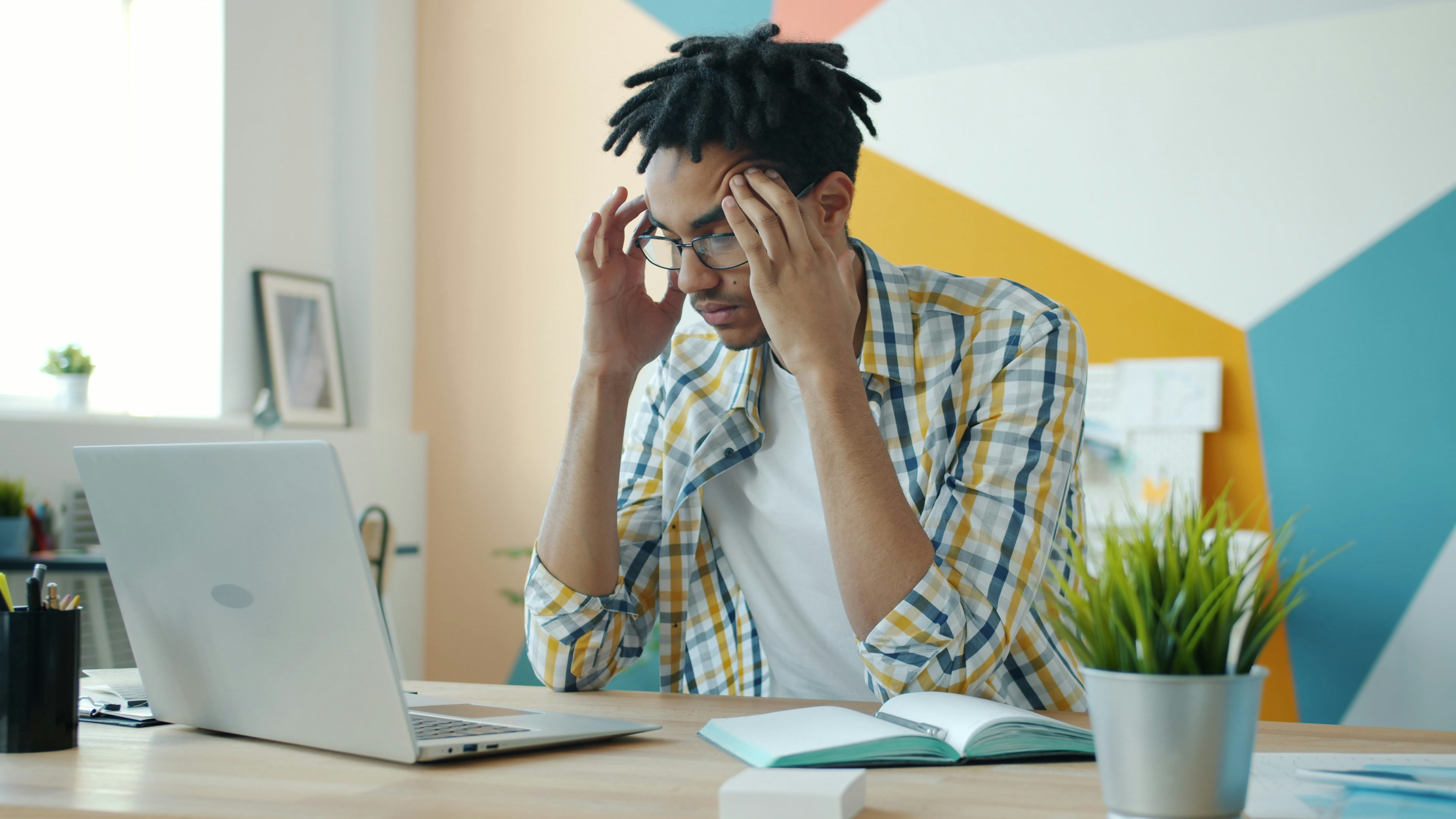 Exhausted mixed race man is feeling headache working with laptop in office room touching head suffering from migraine. People and health concept.