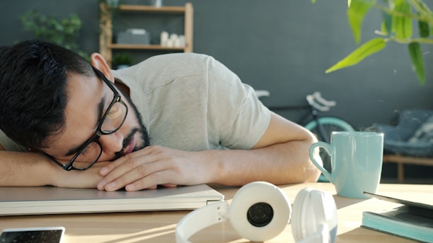 Man sleeping at desk with headphones and coffee.