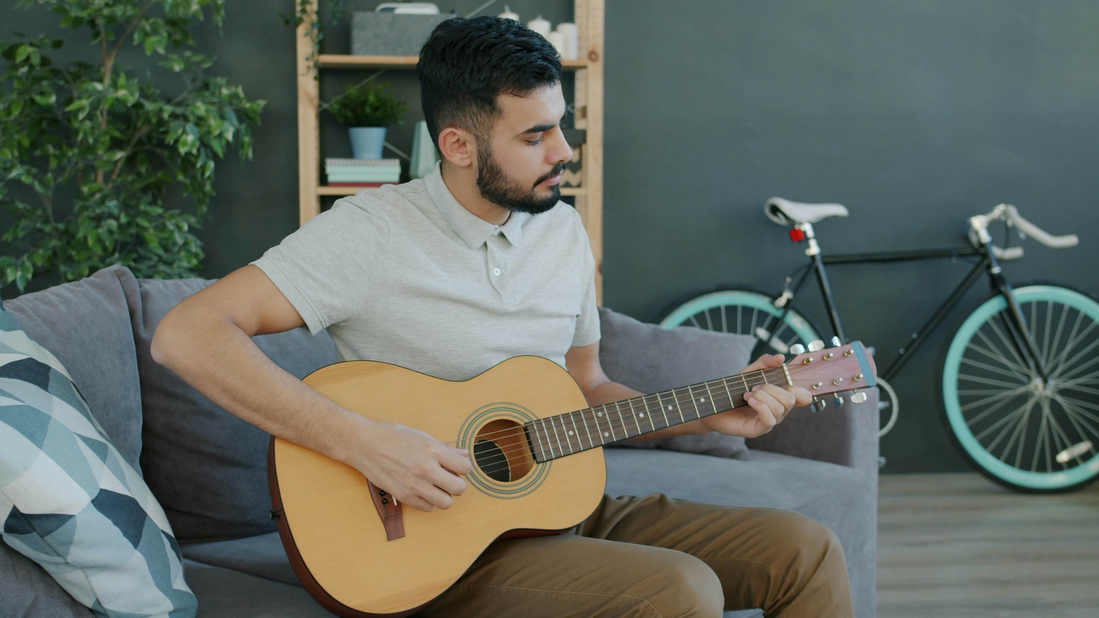 Creative Middle Eastern guy in casual clothing is playing guitar at home enjoying musical instrument sitting on couch alone. People and hobby concept.