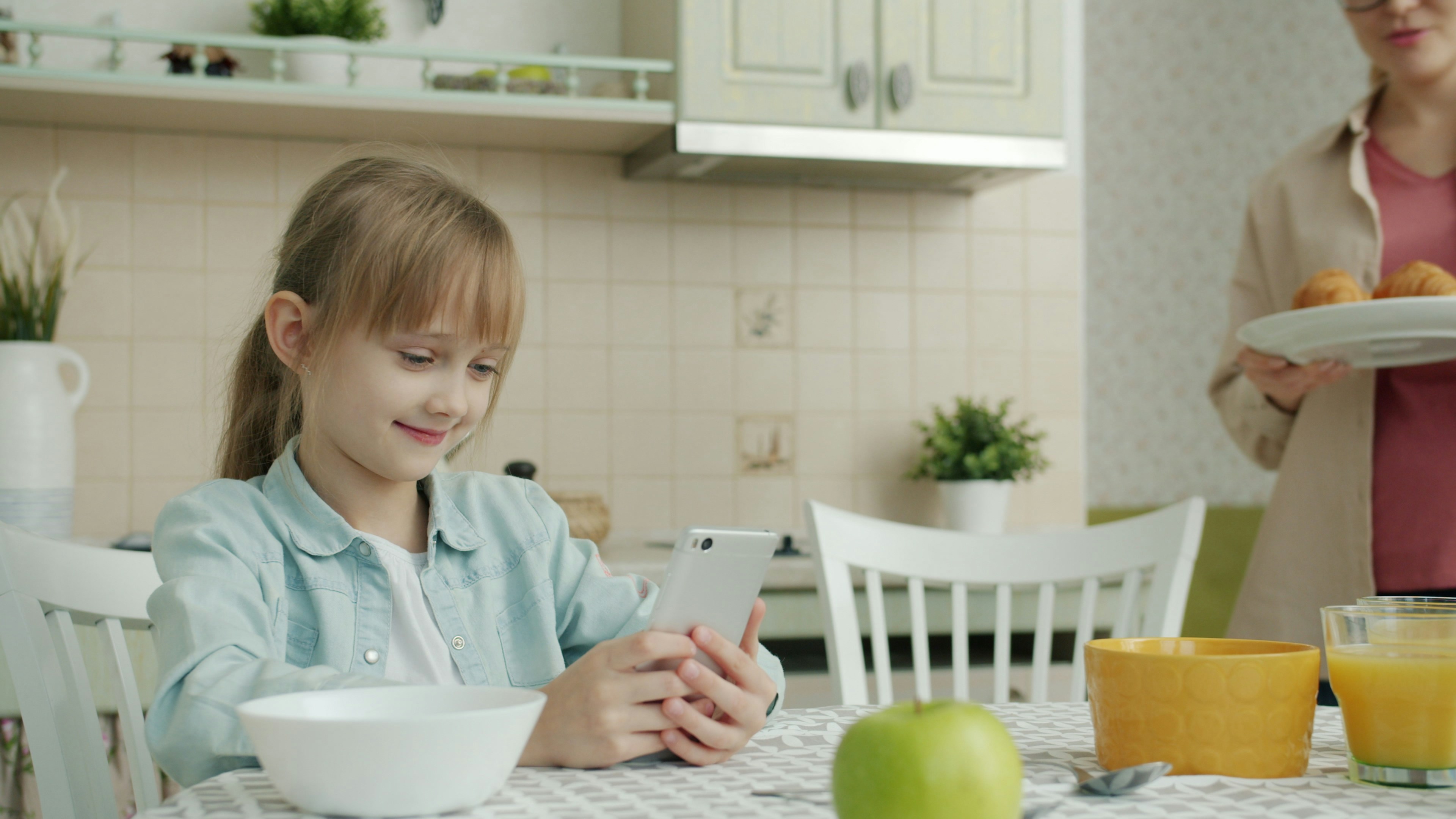 Girl using smartphone while mother brings breakfast
