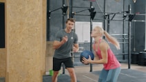 A woman holding a medicine ball with a trainer watching.
